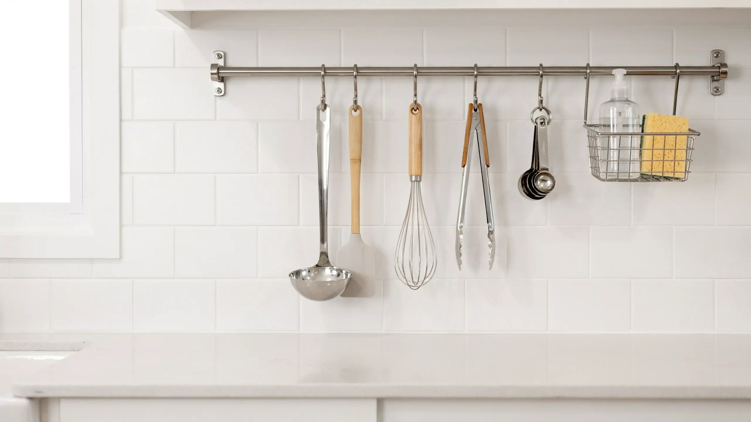 Space-saving stainless steel utensil rail with cooking tools and soap basket, hanging on white tile in a small camper kitchen. 