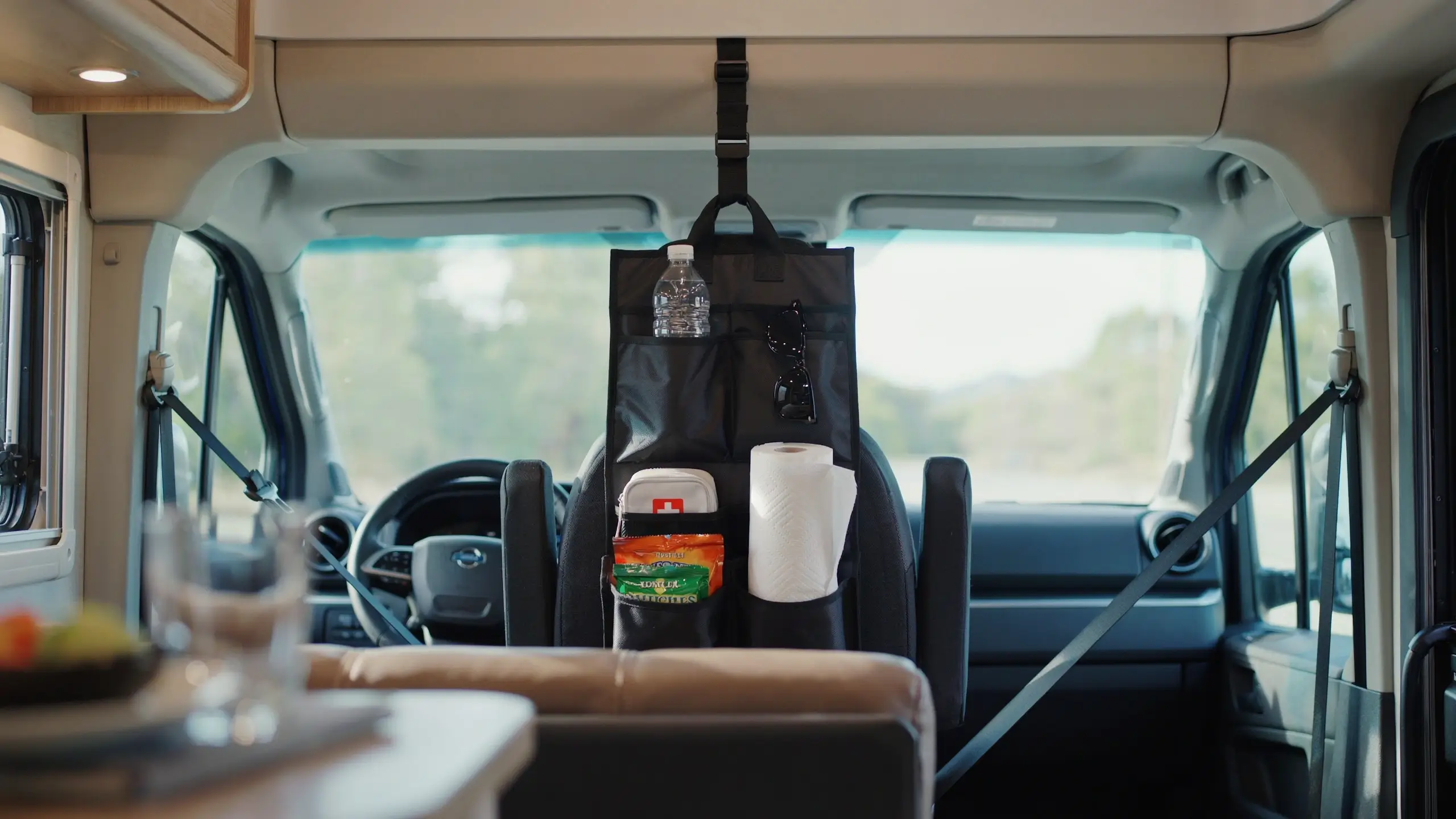 "Black nylon seat-back organizer hanging on the driver's seat of a campervan, holding a plastic water bottle, sunglasses, a white first-aid kit, and a roll of paper towels against a blurred dashboard and nature backdrop."