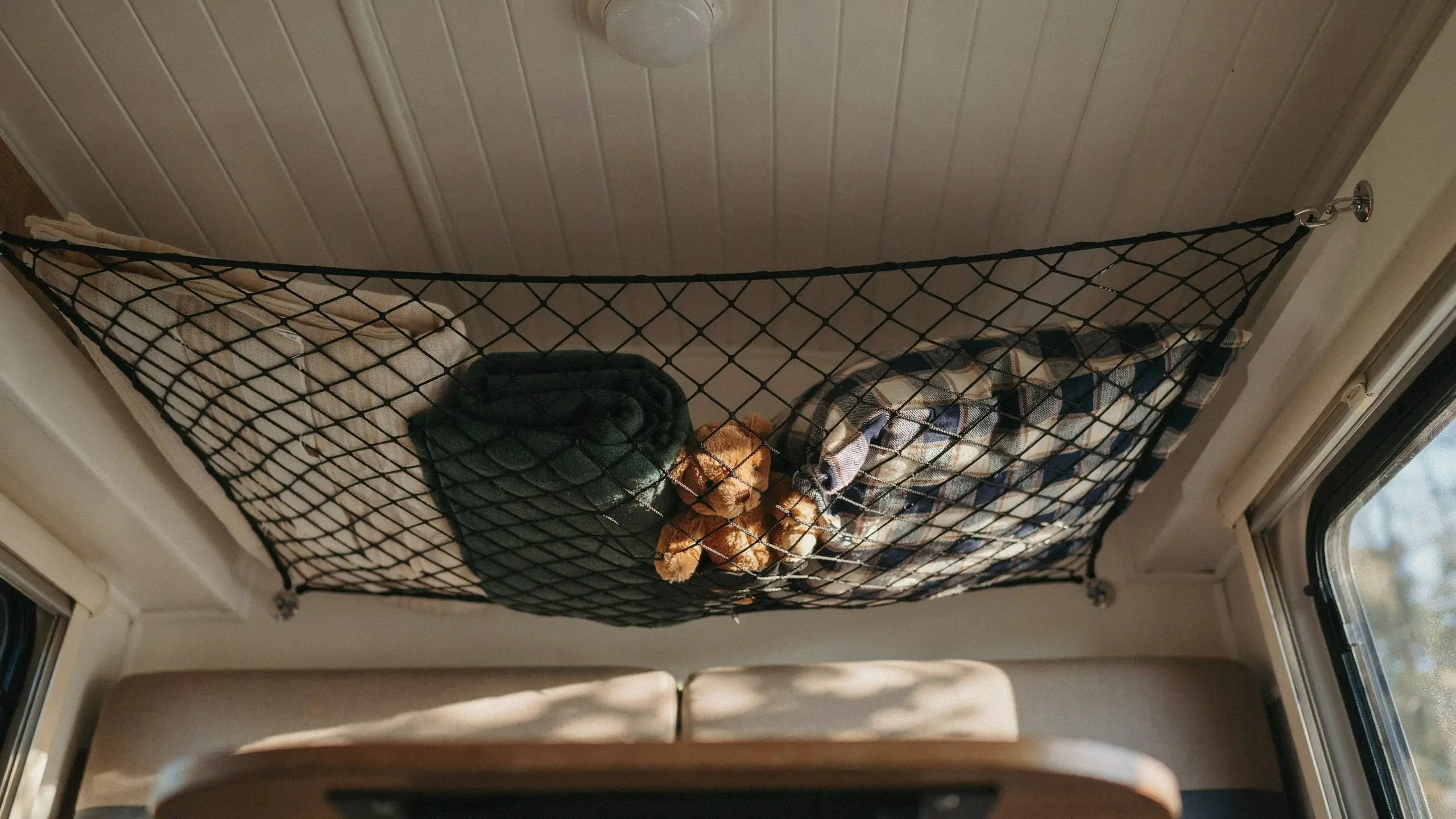 Black mesh ceiling cargo net holding folded beige, green, and plaid blankets along with a small brown teddy bear, installed above the dinette seating in a converted RV camper.