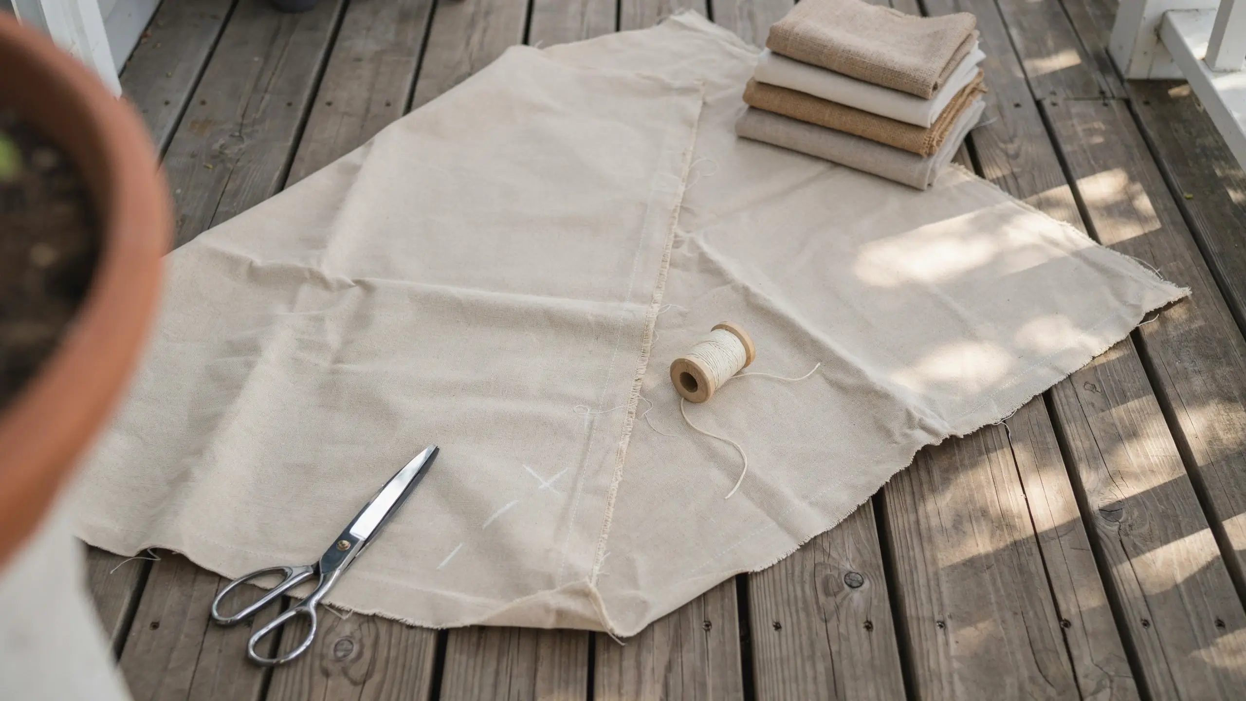 Flat lay of beige natural linen fabric spread across a rustic wooden deck, accompanied by silver scissors, a wooden spool of white thread, and a stack of folded neutral-toned fabric swatches, illustrating how to choose and prep your fabric before starting a sewing project