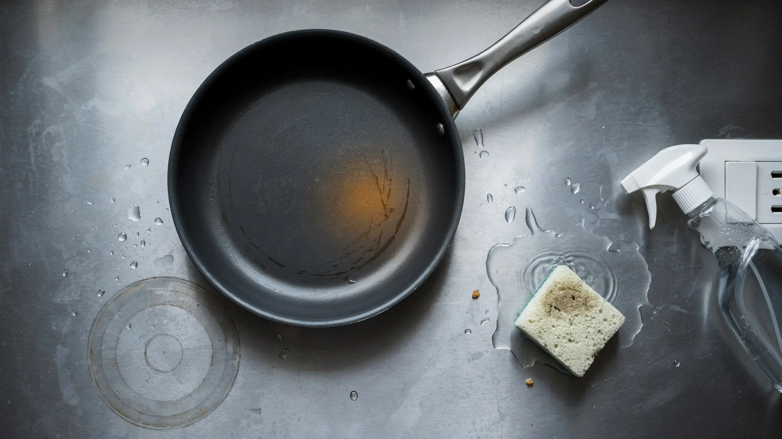 A scratched and burnt non-stick frying pan next to a dirty, soiled sponge and a spray cleaner on a wet stainless steel sink, illustrating common kitchen cleaning mistakes that ruin cookware and waste supplies.