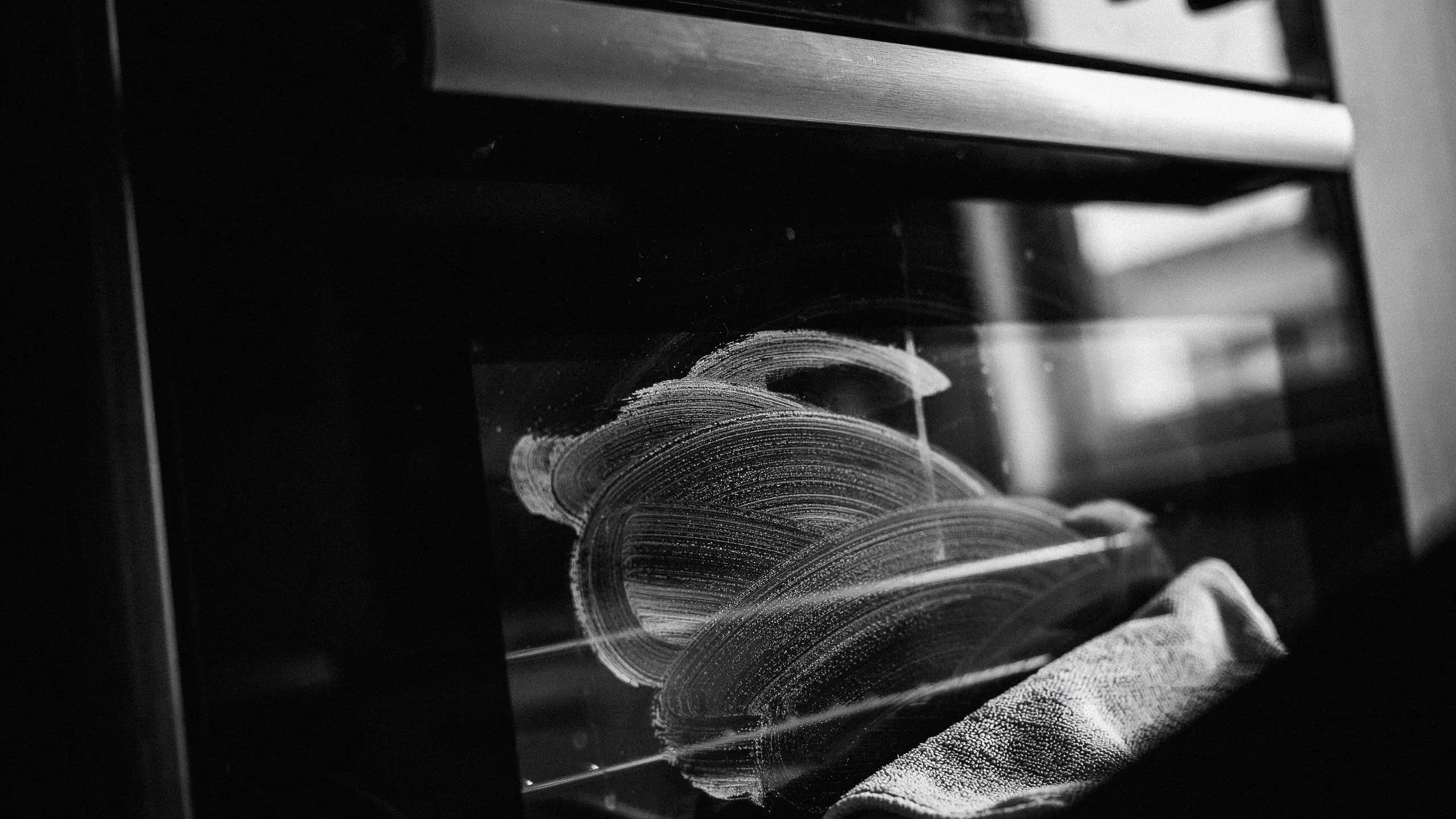Close-up of a dirty oven glass door with circular smudge marks and a textured cleaning cloth.