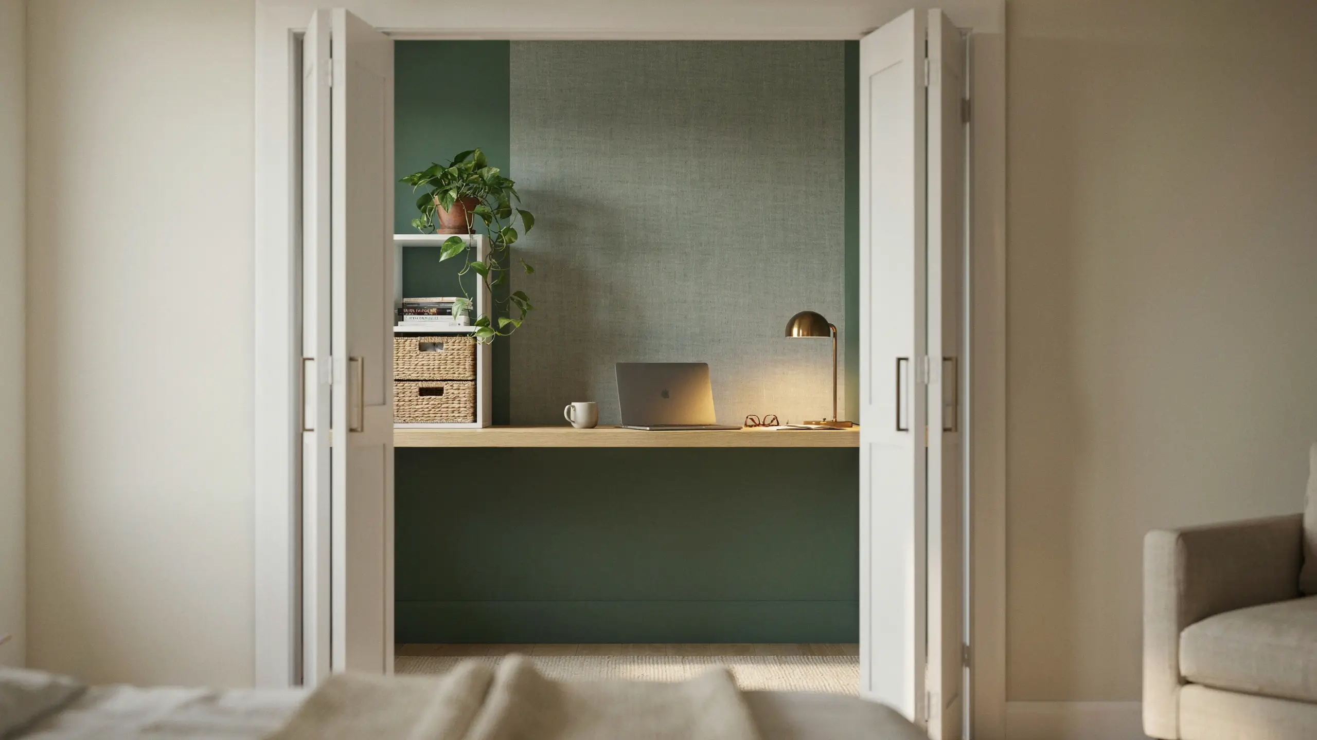 White double doors open to reveal a modern cloffice featuring a floating light-wood desk, an open silver laptop, a glowing brass desk lamp, and a potted trailing plant, all set against a dark sage-green textured accent wall and white shelving with woven baskets. 