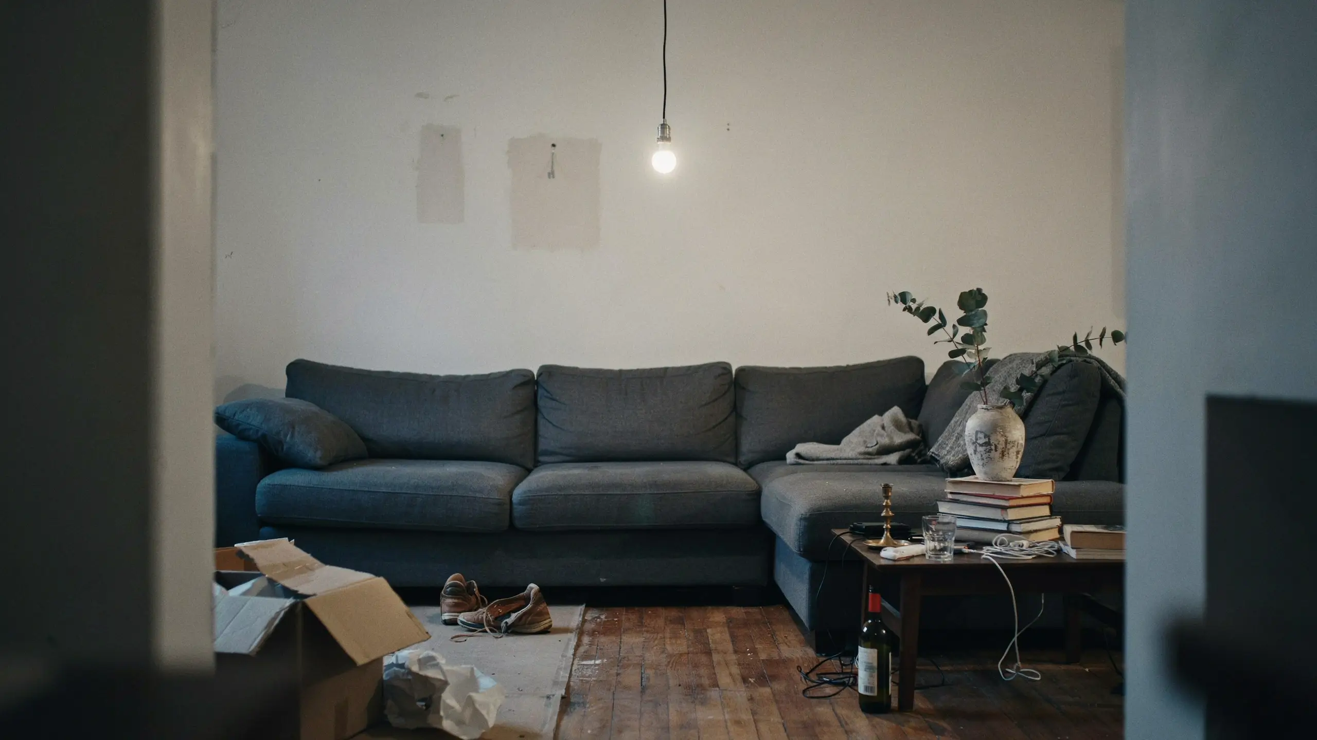 A college apartment living room showing common decorating mistakes like bare walls with patched holes, poor single-bulb lighting, and floor clutter with cardboard boxes and shoes