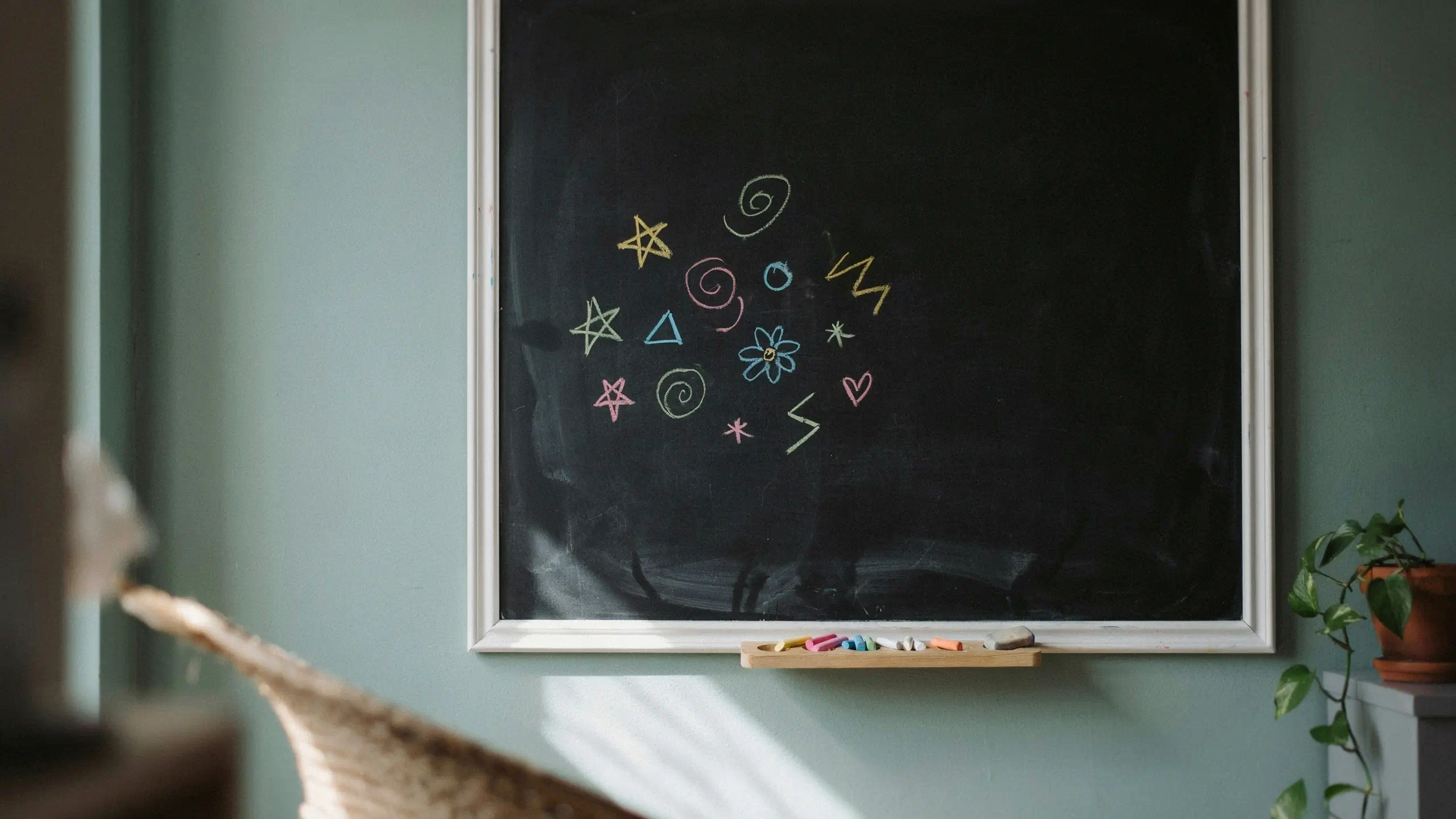 Vibrant chalk drawings of stars, spirals, a flower, and a heart on a black chalkboard mounted on a sage green wall with colorful chalk pieces on the tray. 