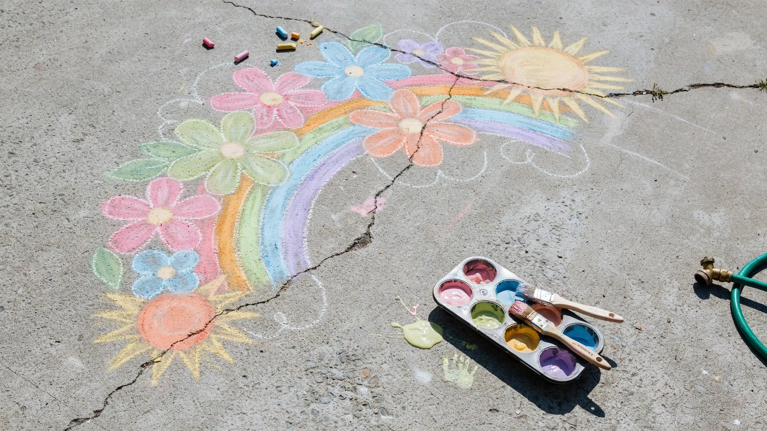 Colorful sidewalk chalk art of a rainbow, flowers, and sun painted outdoors, next to an artist's palette with brushes, scattered chalk, and a garden hose nozzle on cracked gray concrete.