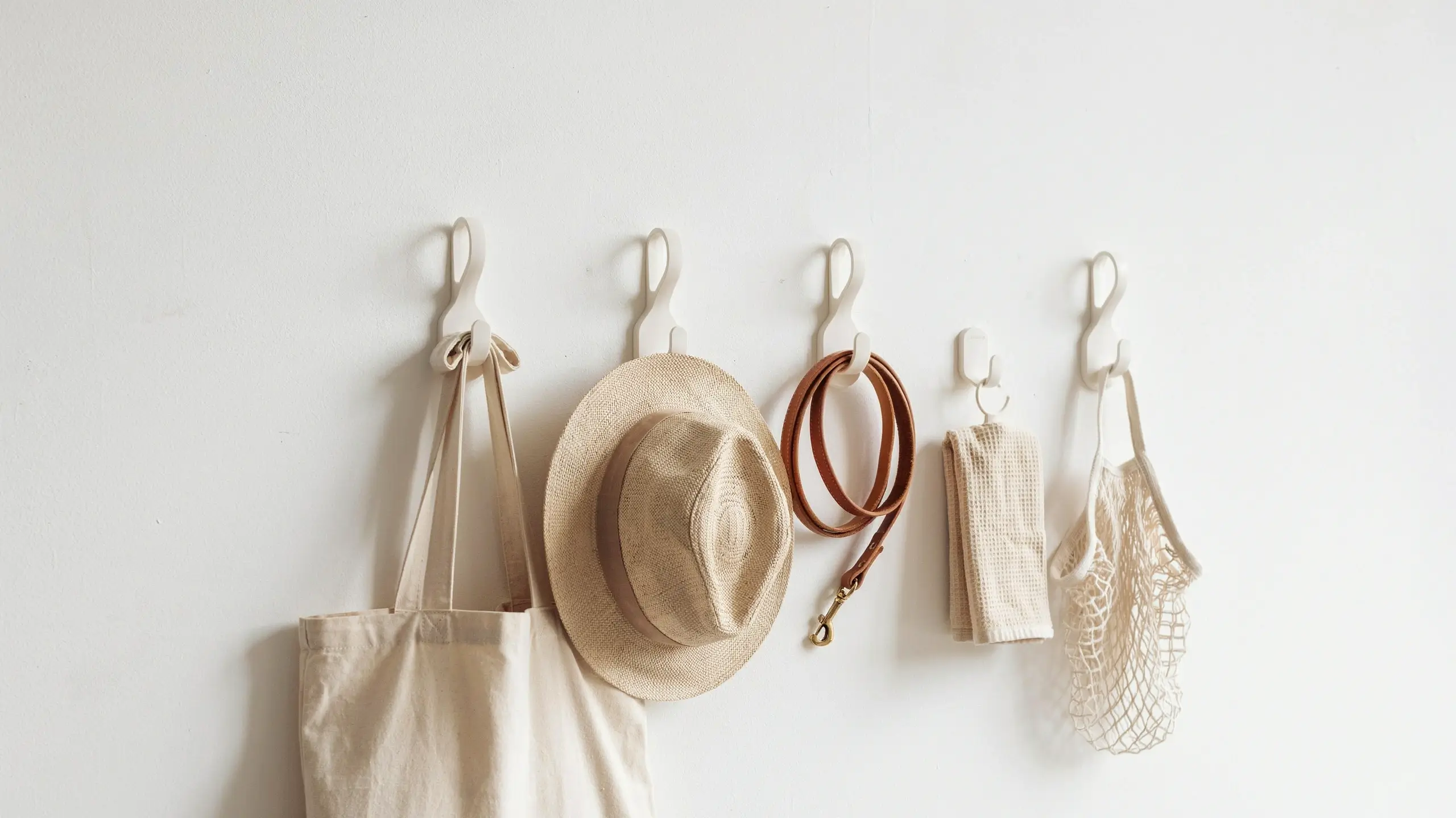 Five white adhesive command hooks hanging a canvas tote bag, woven straw hat, leather dog leash, beige towel, and mesh produce bag on a plain white entryway wall. 
