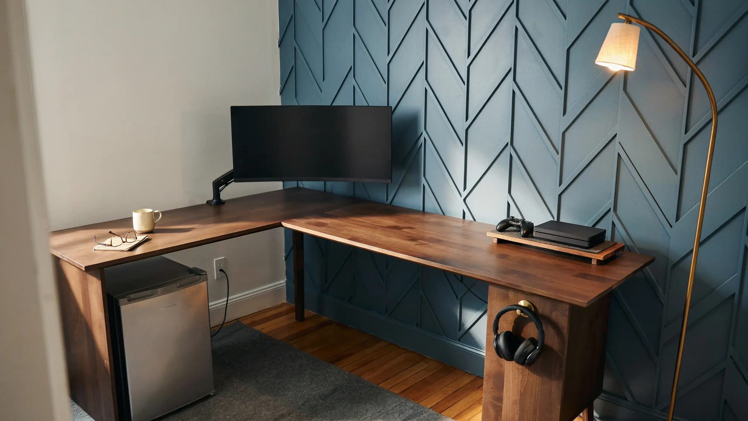 L-shaped corner desk gaming setup in a spare bedroom mini cave with a curved monitor, console, and mini fridge. 