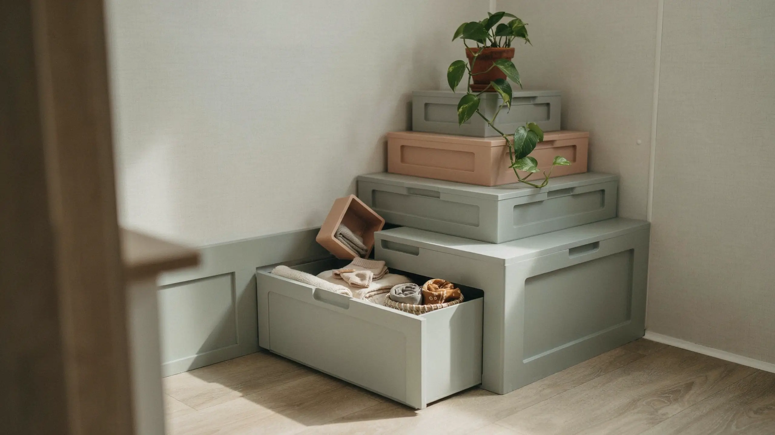 A stack of sage green and dusty pink modular storage bins arranged in a step-like tower to utilize an awkward room corner, topped with a trailing green pothos plant, with the bottom drawer open to reveal neatly folded linens and a small woven basket.