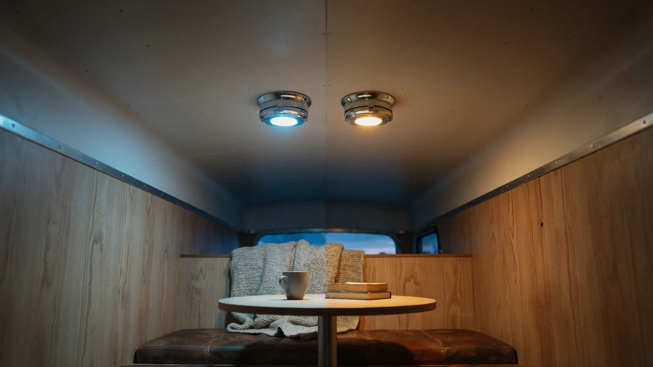 A cozy campervan interior with wooden paneling and a round table, showing a cool blue overhead light on the left and a warm yellow bulb installed on the right to create a snug evening atmosphere