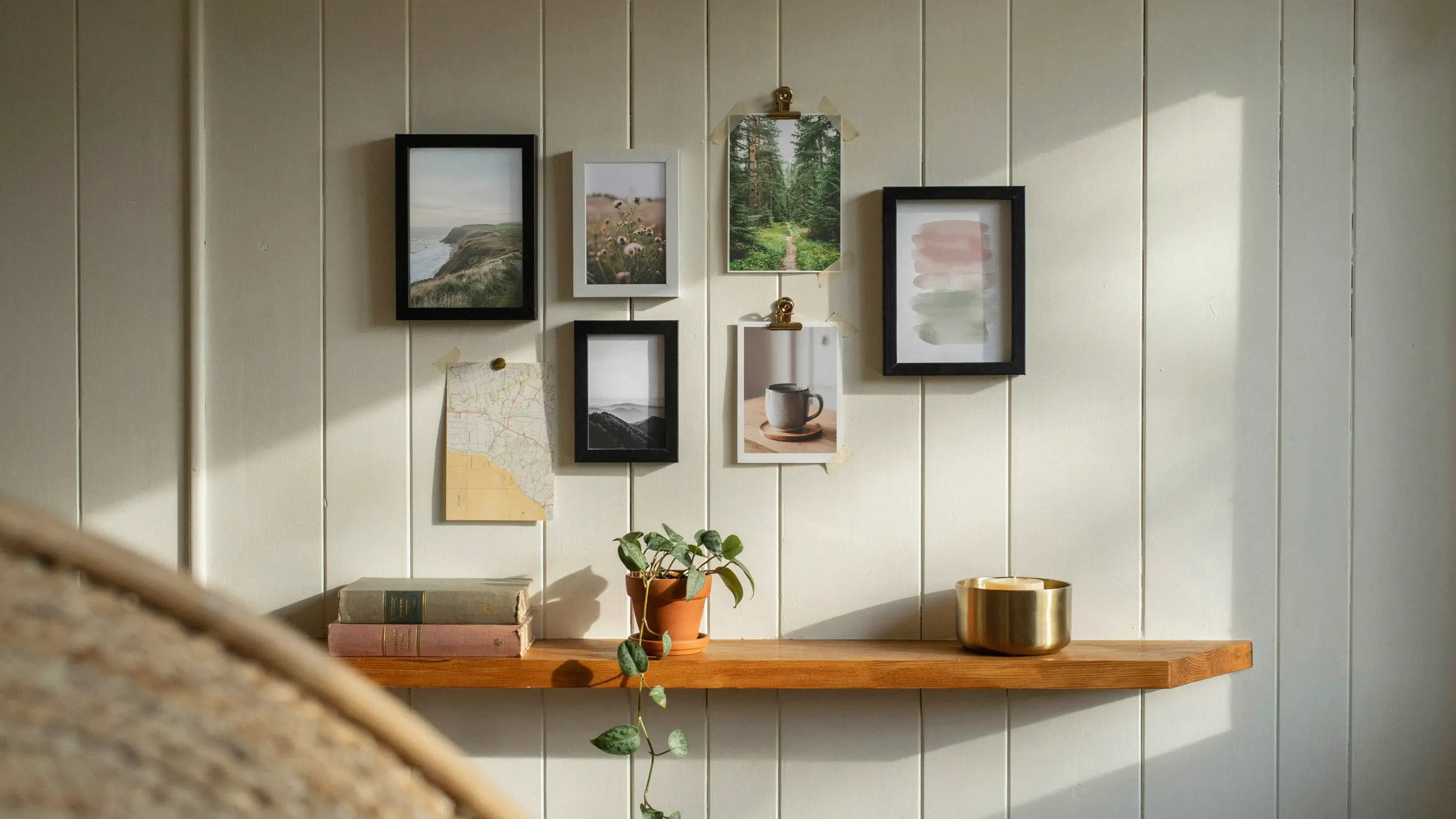 White vertical wood paneling featuring a wooden floating shelf styled with a stack of vintage books, a terracotta pot with a trailing ivy plant, a gold candle, and a gallery wall of framed landscape art. 