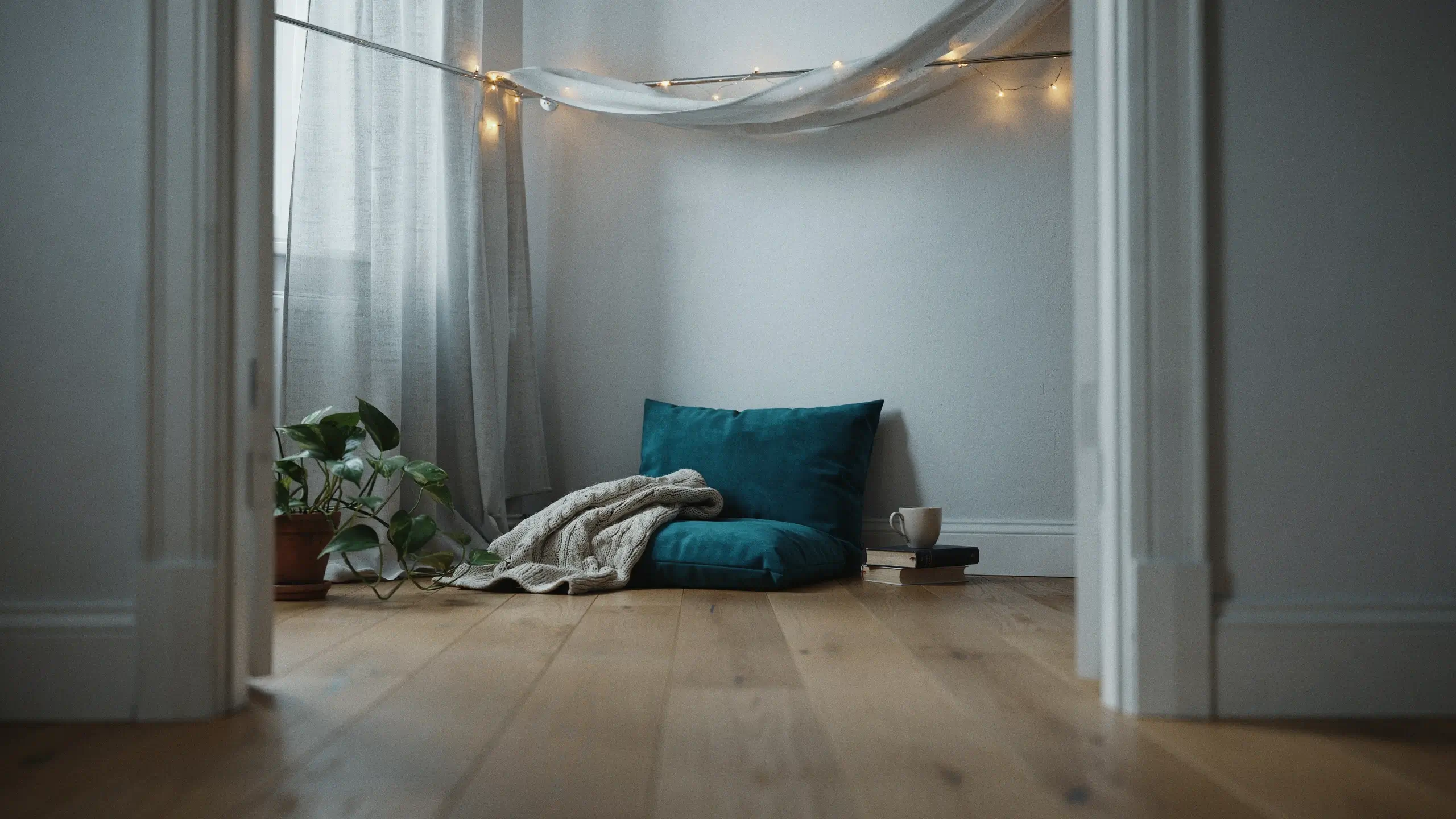A cozy floor seating hideaway with a plush teal cushion, a cream chunky-knit blanket, warm string lights, and a terracotta potted plant placed beside a sheer white curtain and window. 