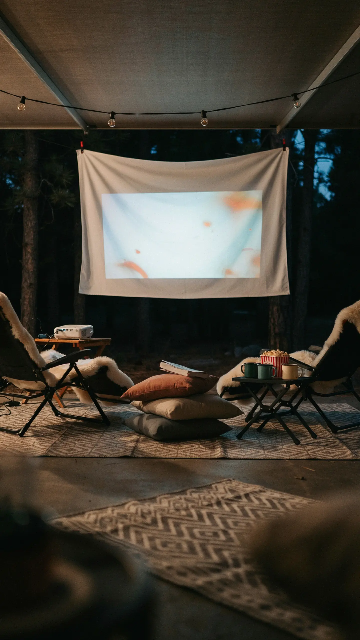 Cozy outdoor movie setup under a canopy awning featuring a projector, white screen, warm string lights, fur-lined chairs, and floor pillows against a dark pine forest at dusk. 