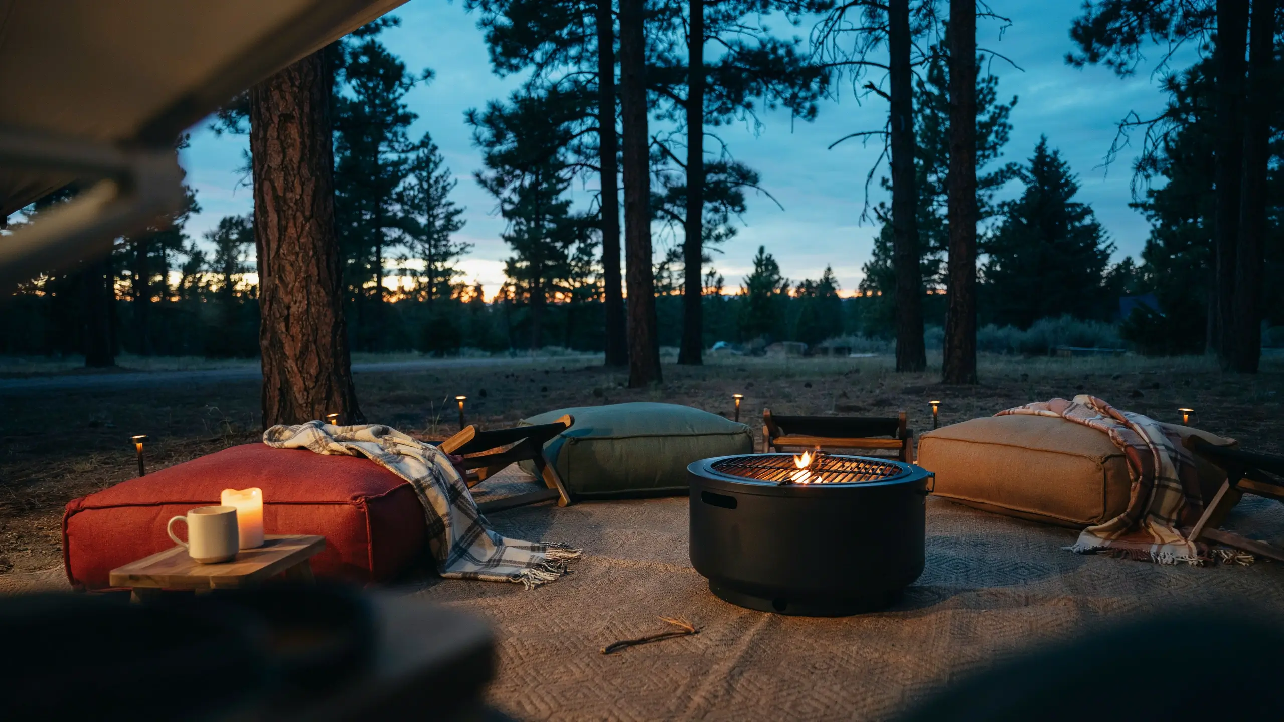 A cozy twilight camping scene featuring large red, olive, and tan floor cushions arranged around a black portable fire pit on a textured outdoor rug, with a white ceramic mug and candle on a small wooden tray, set within a pine forest under a dramatic sunset sky."