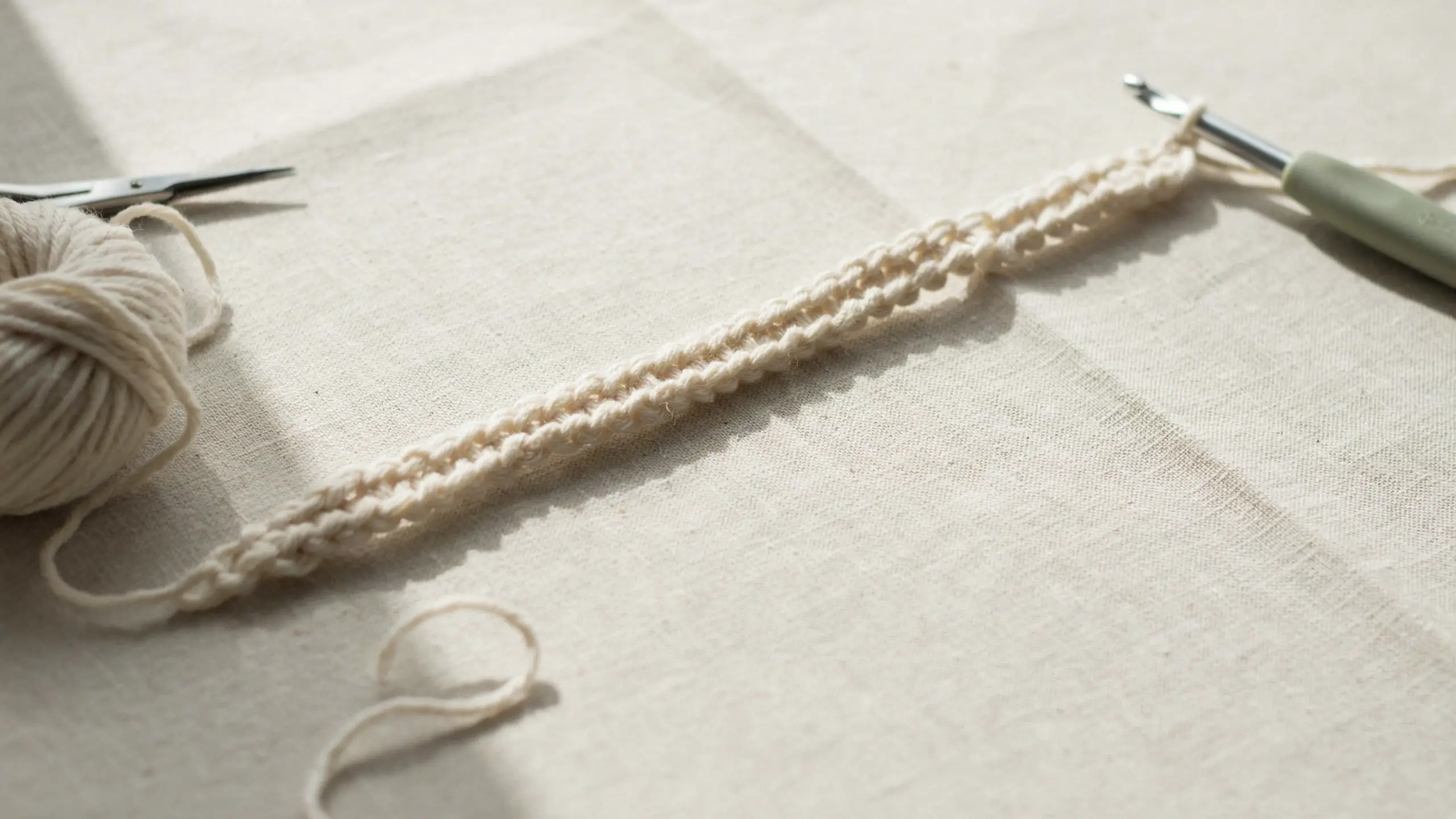 Close-up flat lay of a cream-colored cotton crochet chain stitch foundation row, featuring a sage green ergonomic crochet hook and a ball of yarn, illuminated by soft natural sunlight with distinct shadows on a textured linen cloth.