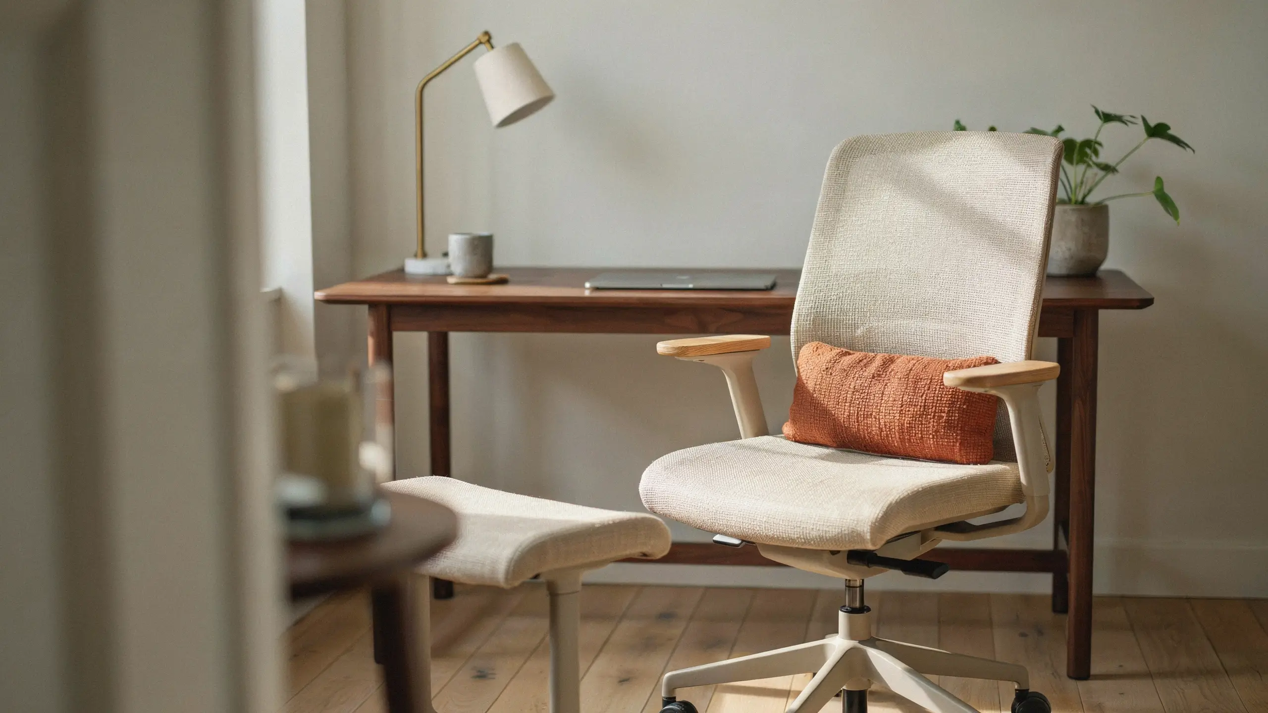 Modern cream ergonomic office chair with wooden armrests and an orange lumbar pillow in a sunlit home office, paired with a dark wood desk, brass lamp, and potted plant. 