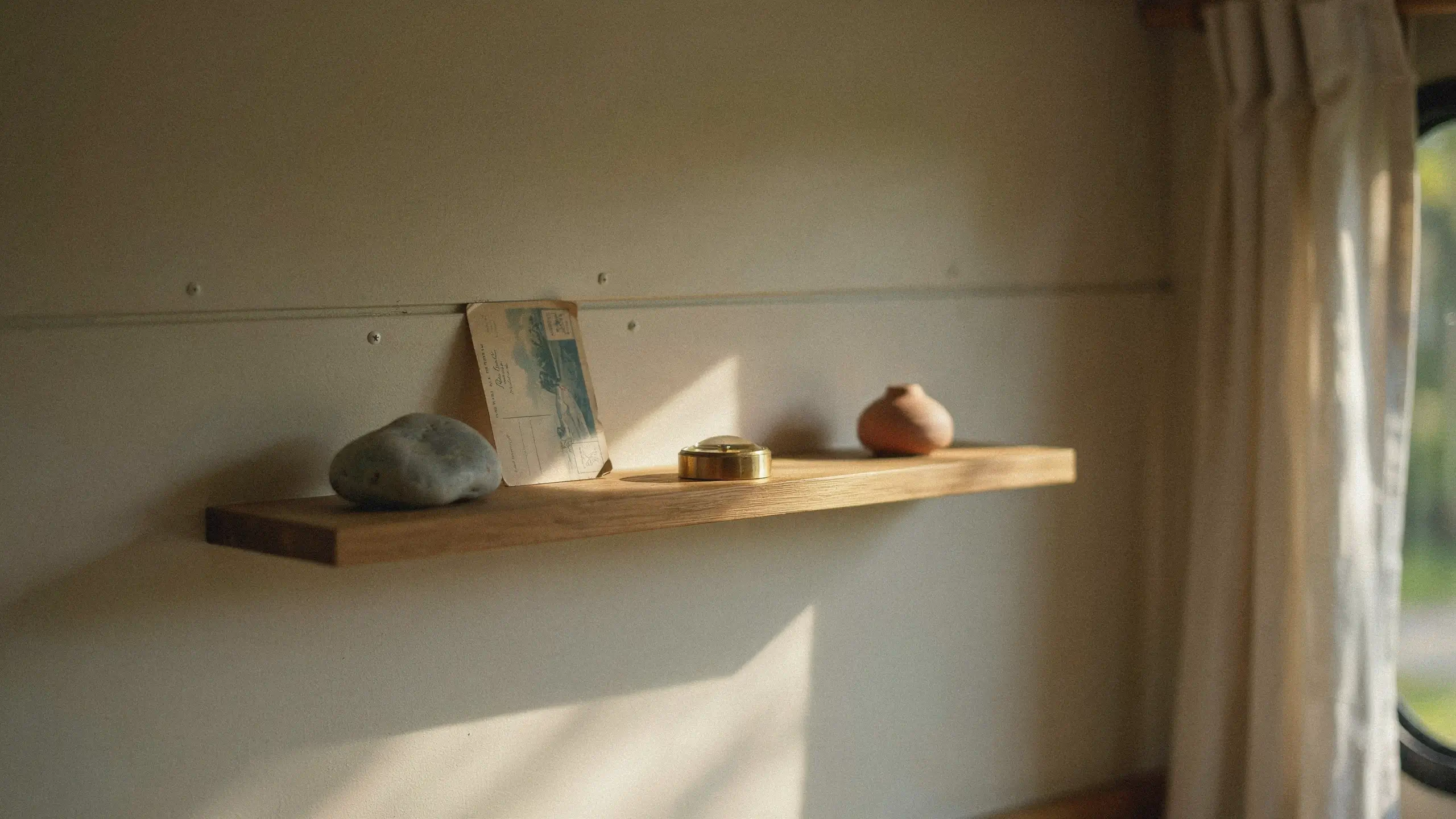 A minimalist floating wooden shelf holding a grey river stone, vintage coastal postcard, brass lidded box, and small terracotta vase, illuminated by diagonal warm sunlight. 