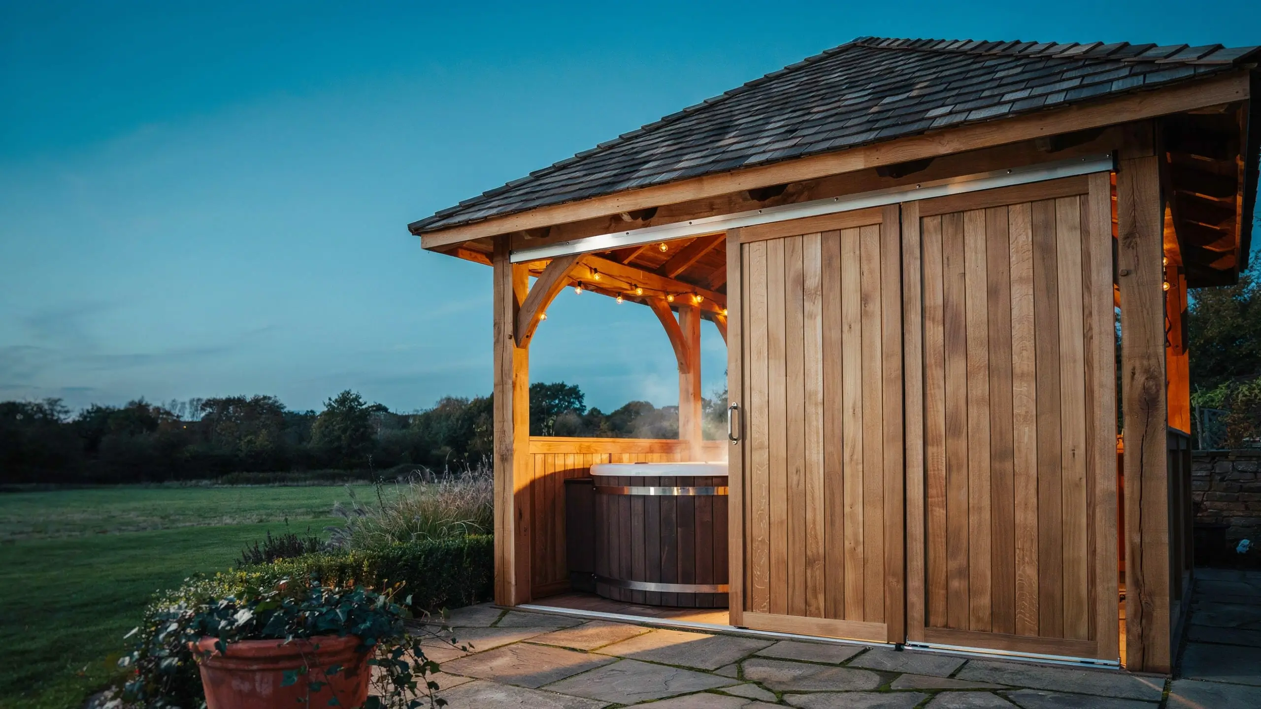 Wooden hot tub gazebo with sliding panel doors, glowing string lights, and steam rising into a twilight evening sky.