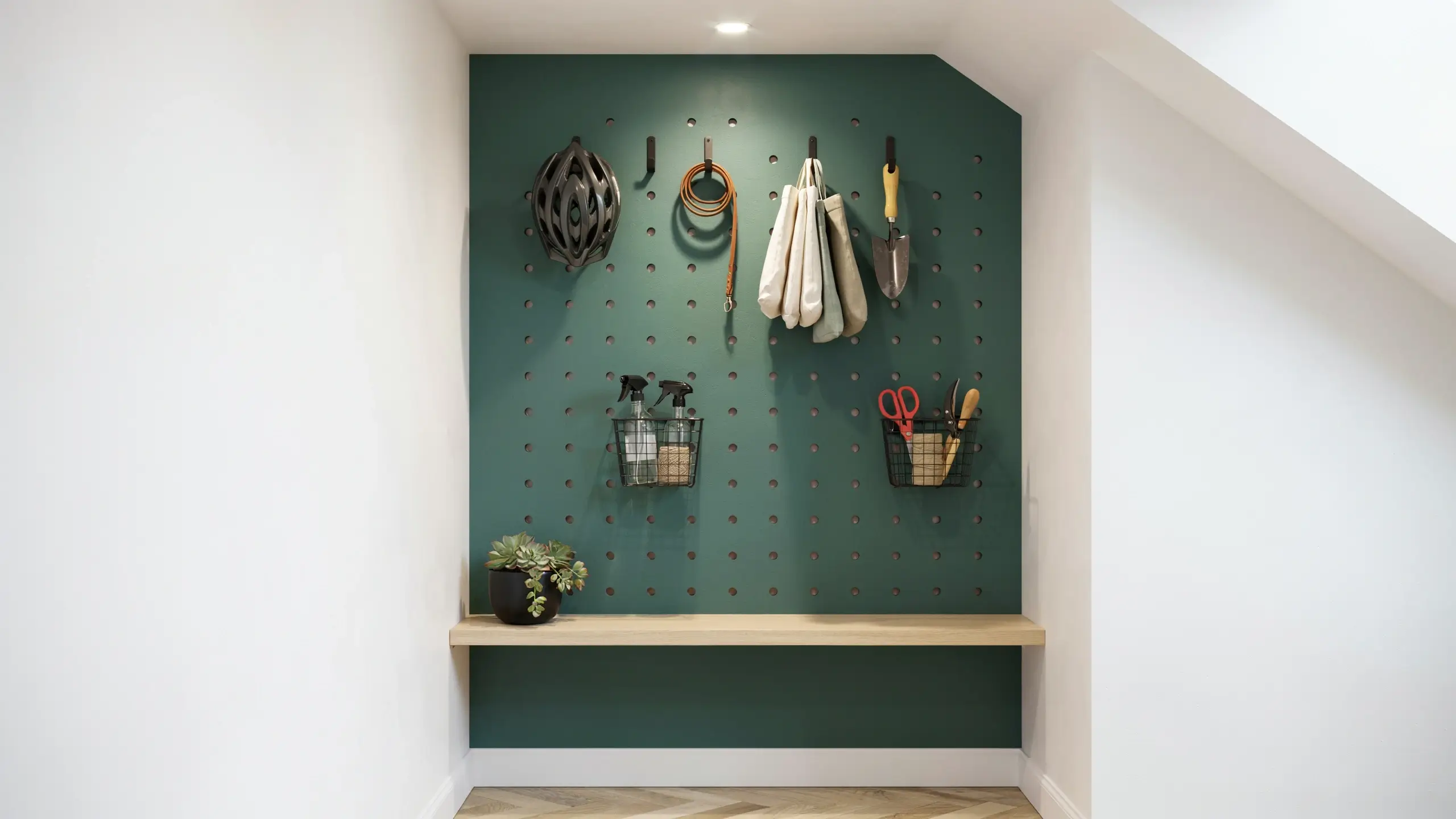 A dark forest green pegboard accent wall fitted with black hooks and wire baskets, organizing gardening tools, spray bottles, a dog leash, and a bicycle helmet in a sloped attic nook, topped with a light wood floating shelf holding a potted succulent plant.
