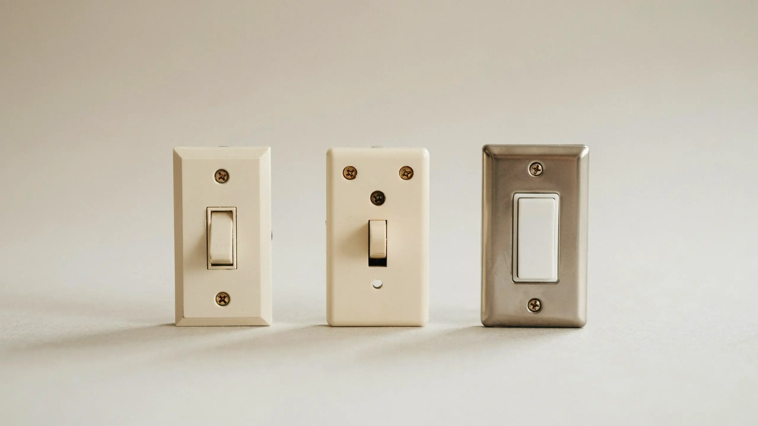 Three light switch types displayed in a row on a beige background: two traditional beige toggle switches and a white Decora rocker switch with a brushed metal wall plate.