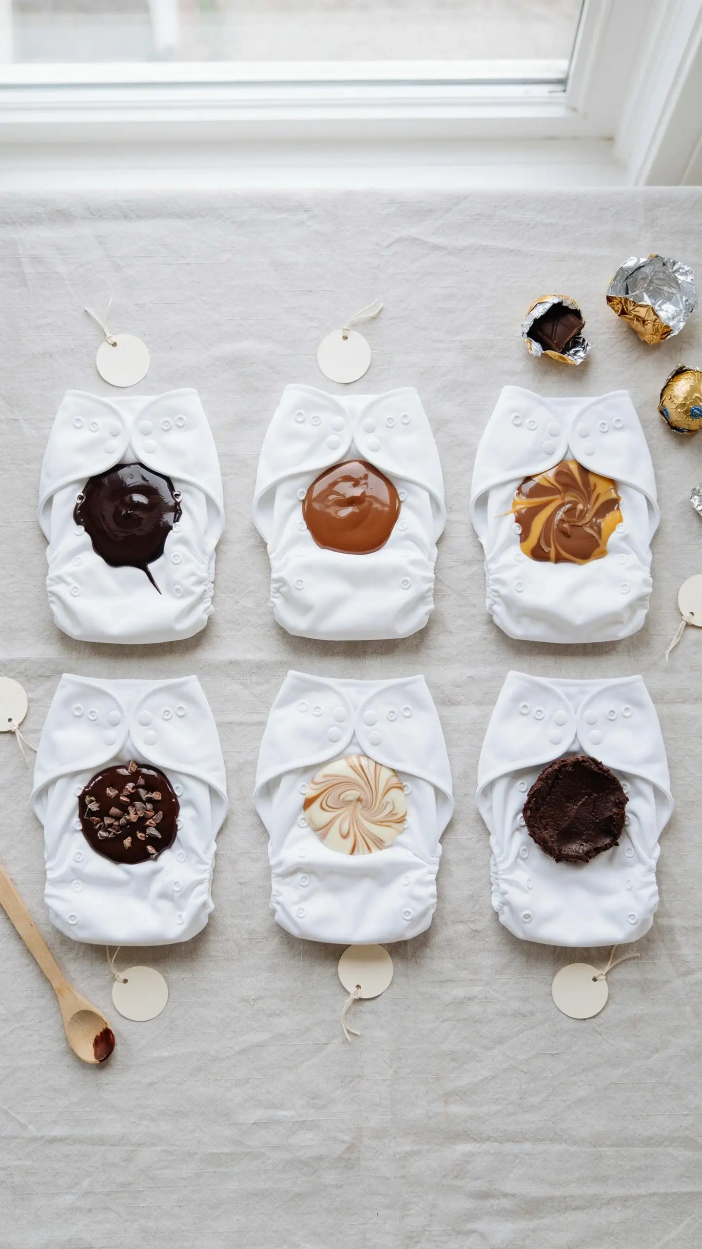 Six white reusable cloth diapers laid out on a white table setup for a baby shower dirty diaper game, featuring unique chocolate, caramel, and marbled fudge dollops that look like messy diapers, alongside empty foil wrappers and a wooden spoon. 