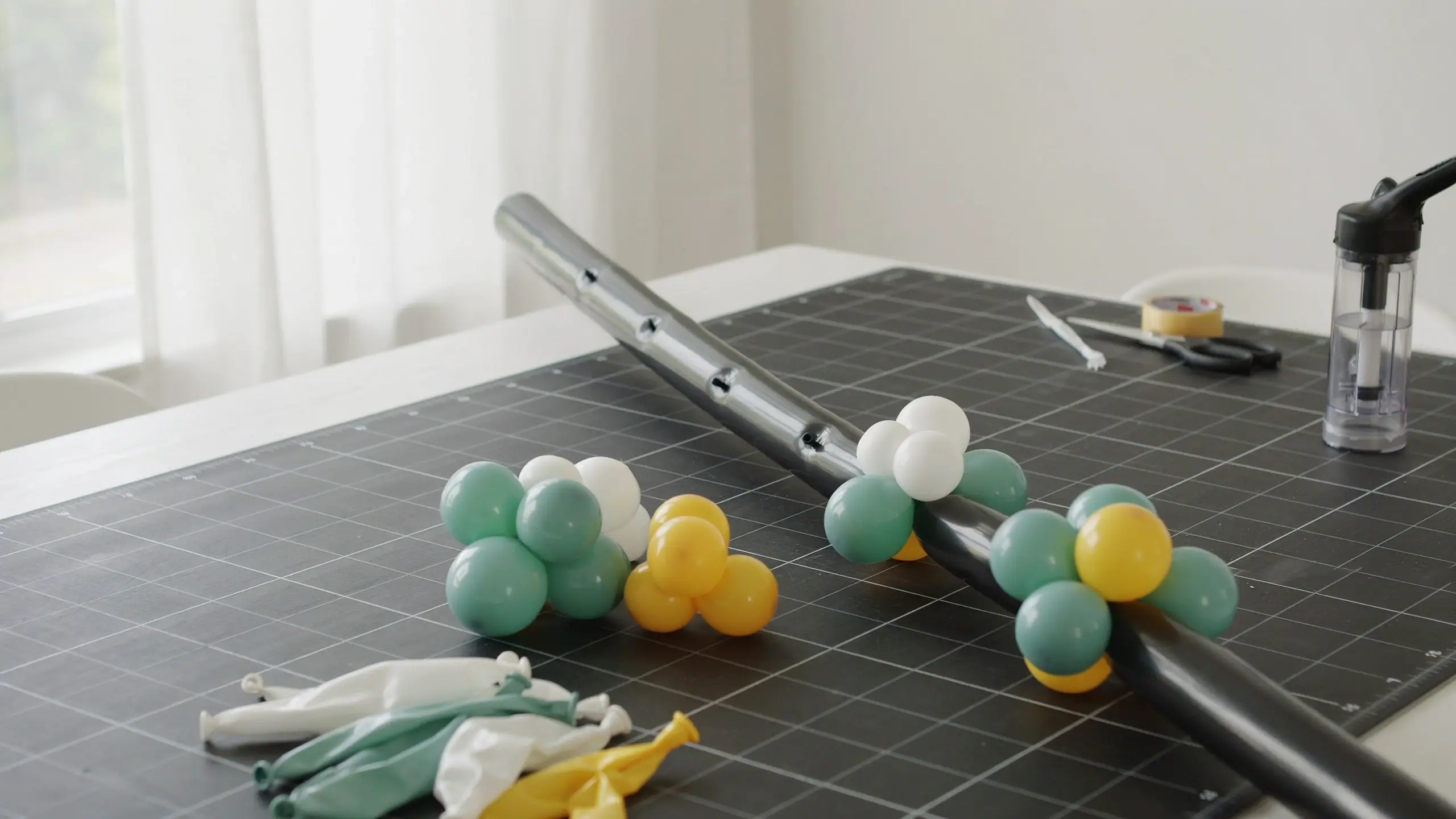 Step-by-step DIY balloon arch tutorial step 5: threading small clusters of sage green, white, and yellow balloons onto a plastic balloon arch strip on a crafting table with tools