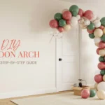 A DIY balloon arch in sage green, dusty rose, and cream colors installed next to a white door in a modern living room, with a balloon pump and supplies on the floor, illustrating an easy step-by-step party decoration tutorial