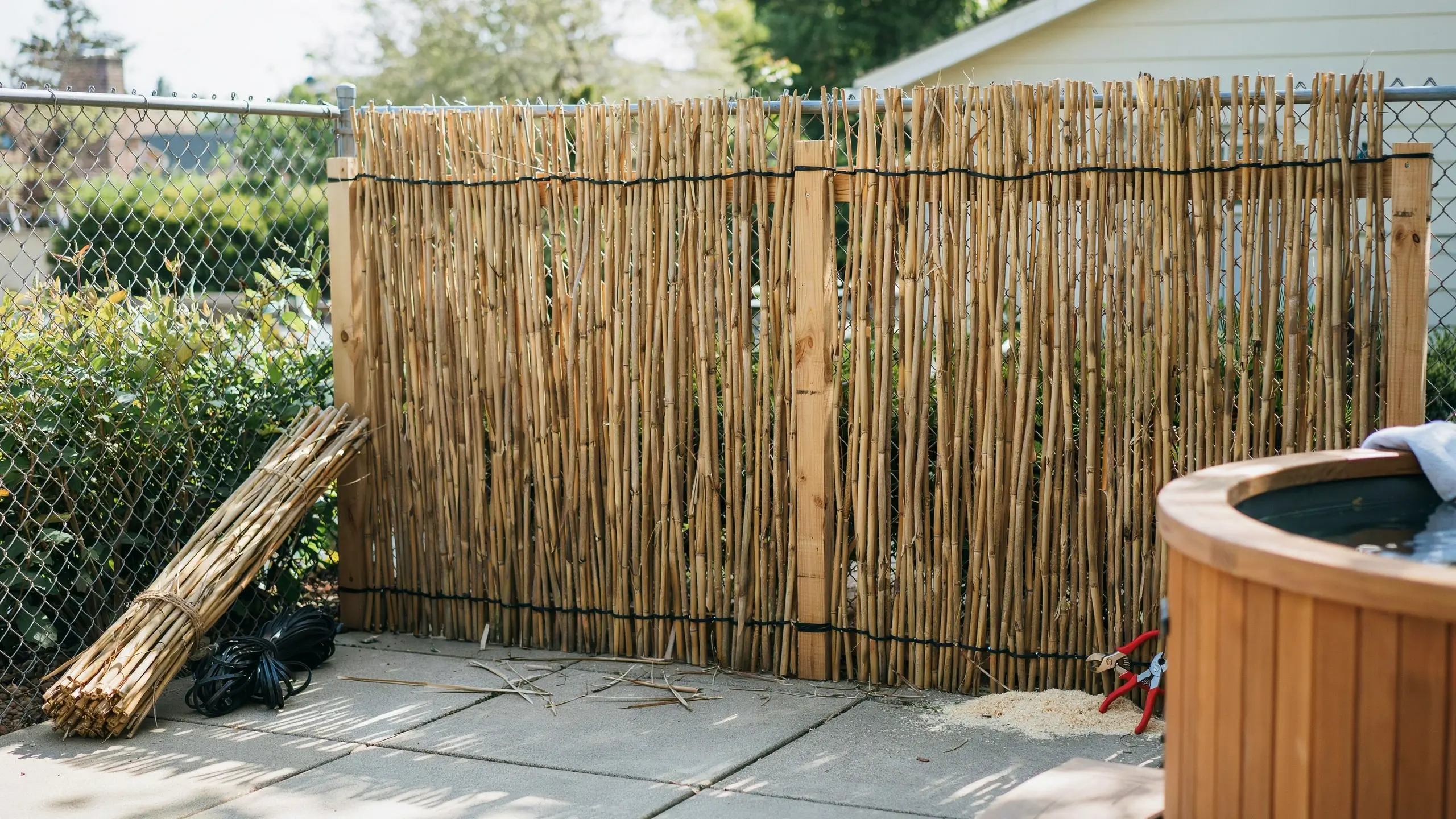 A close-up view of a completed DIY bamboo reed privacy screen installed vertically over a metal chain-link fence. The lightweight reeds are secured with horizontal black ties and wooden support posts. A spare bundle of bamboo reeds, a pile of black fasteners, and a pair of red-handled wire cutters sit on the concrete backyard patio, next to a wooden hot tub.