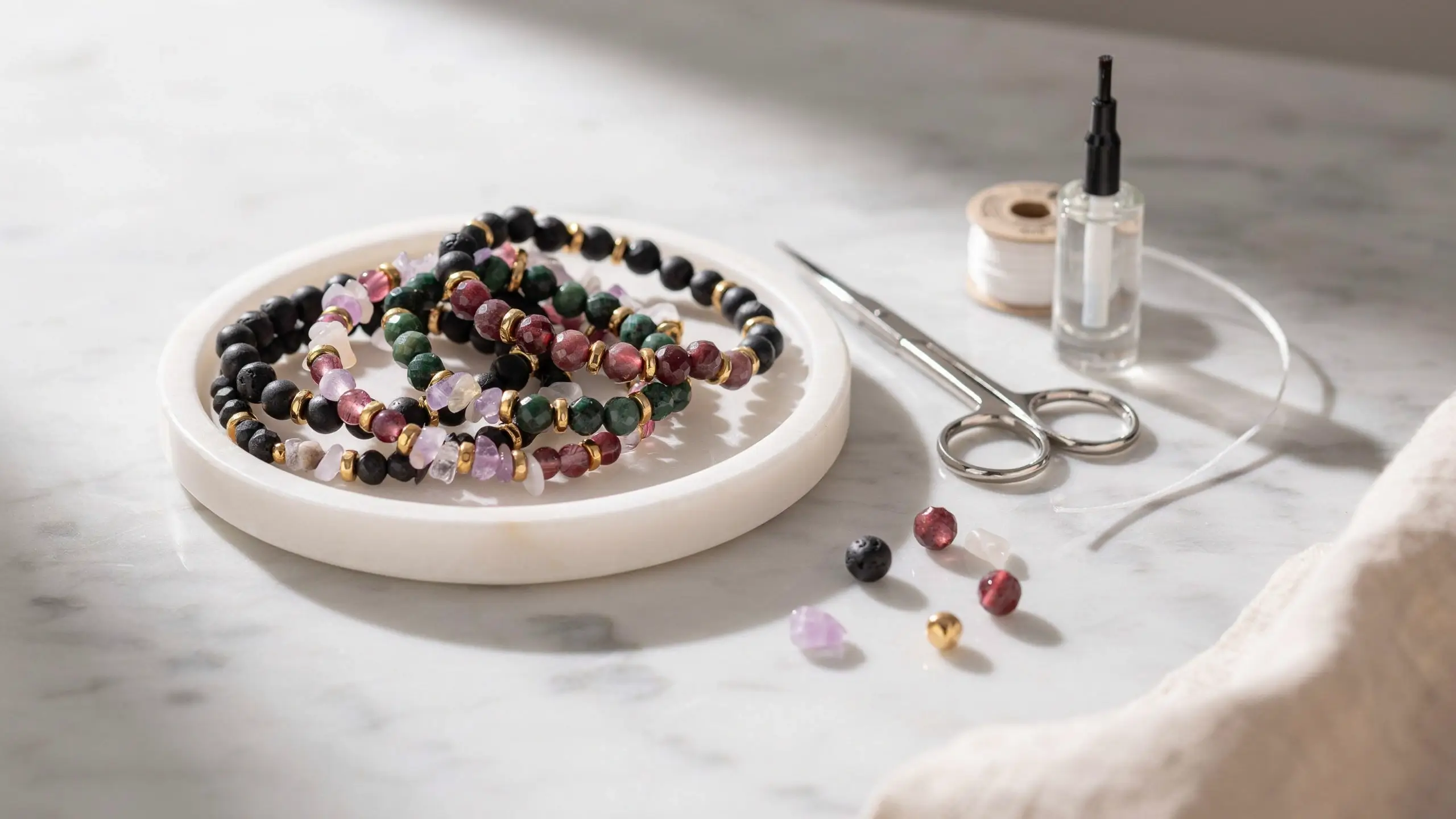Handmade chunky bead and seed bead bracelets on a white tray with scissors, elastic cord, and glue for DIY jewelry making on a marble surface. 
