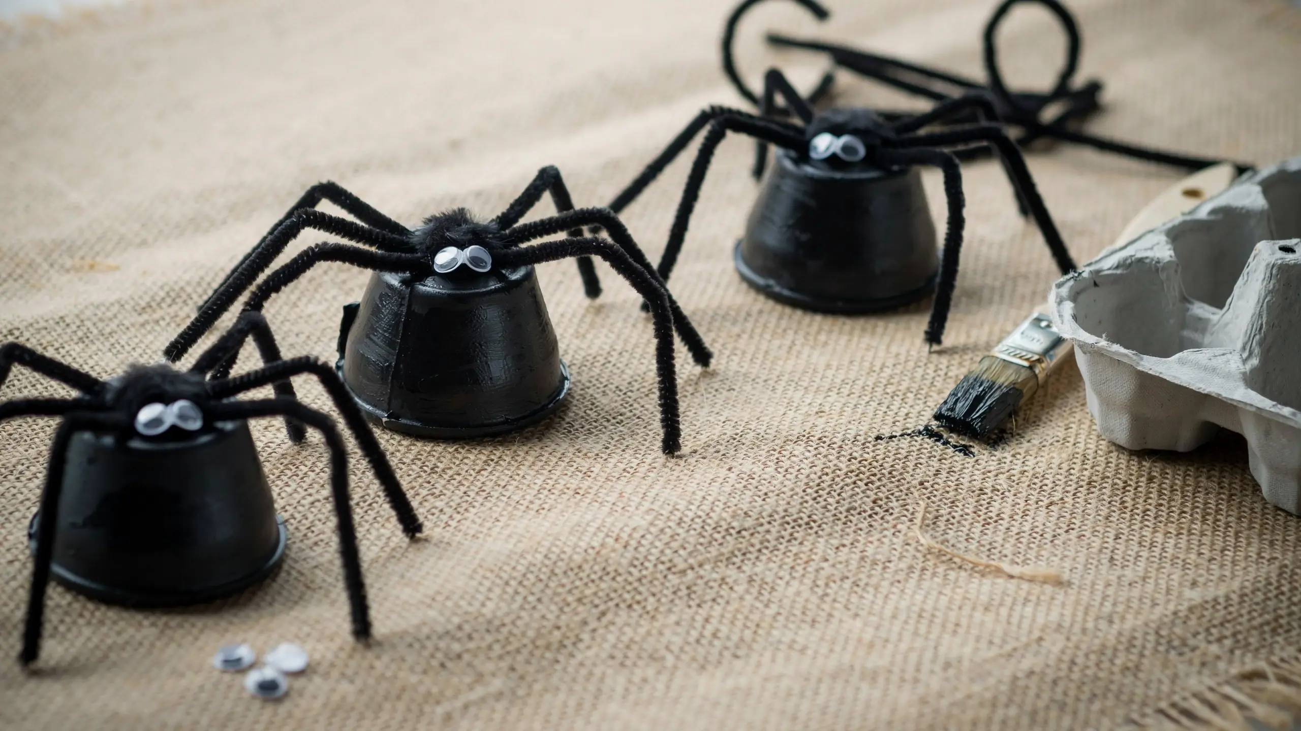 Three DIY black spiders made from painted egg carton cups with fuzzy pipe cleaner legs and googly eyes sitting on burlap with crafting supplies nearby.