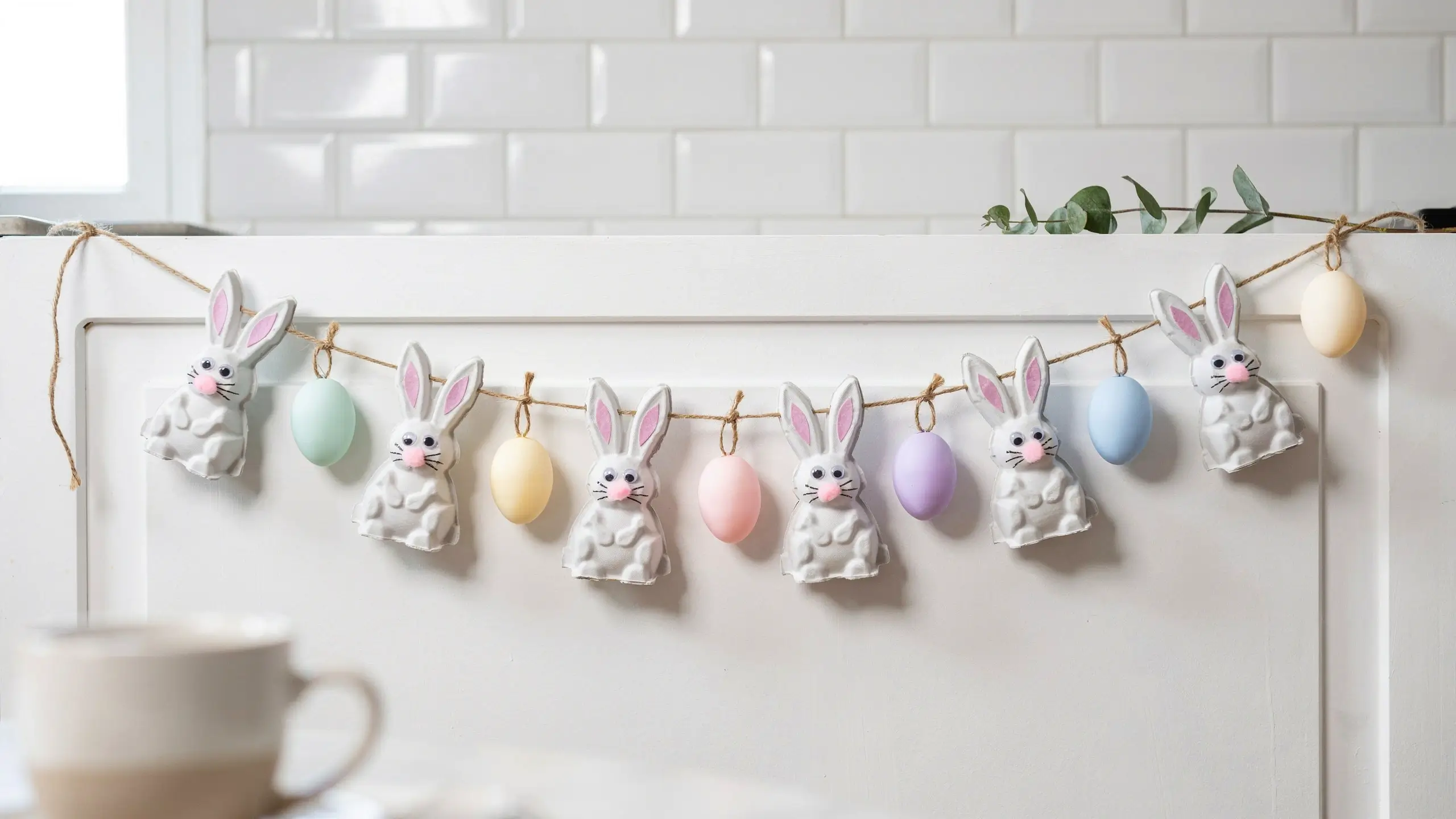 A handmade DIY Easter garland made from upcycled white egg carton bunnies with pink ears and pastel colored eggs, hanging on natural twine against a white kitchen cabinet and subway tile backsplash. 