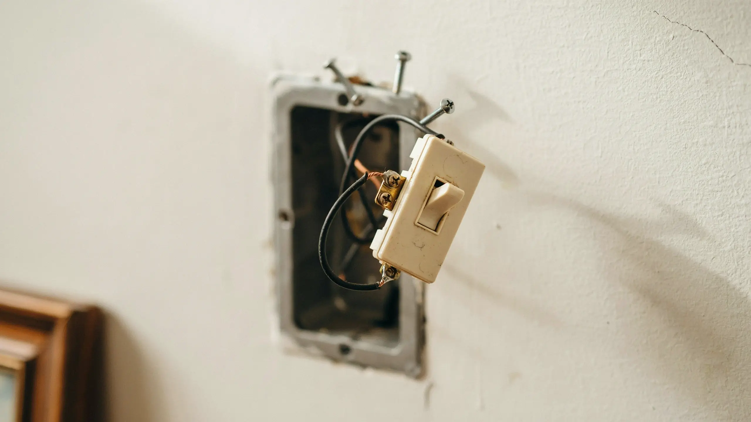 An off-white toggle light switch dangling out of a metal wall box, showing exposed copper ground wire and black electrical wires attached to brass terminal screws, captured during Step 3 of a home improvement wiring replacement.