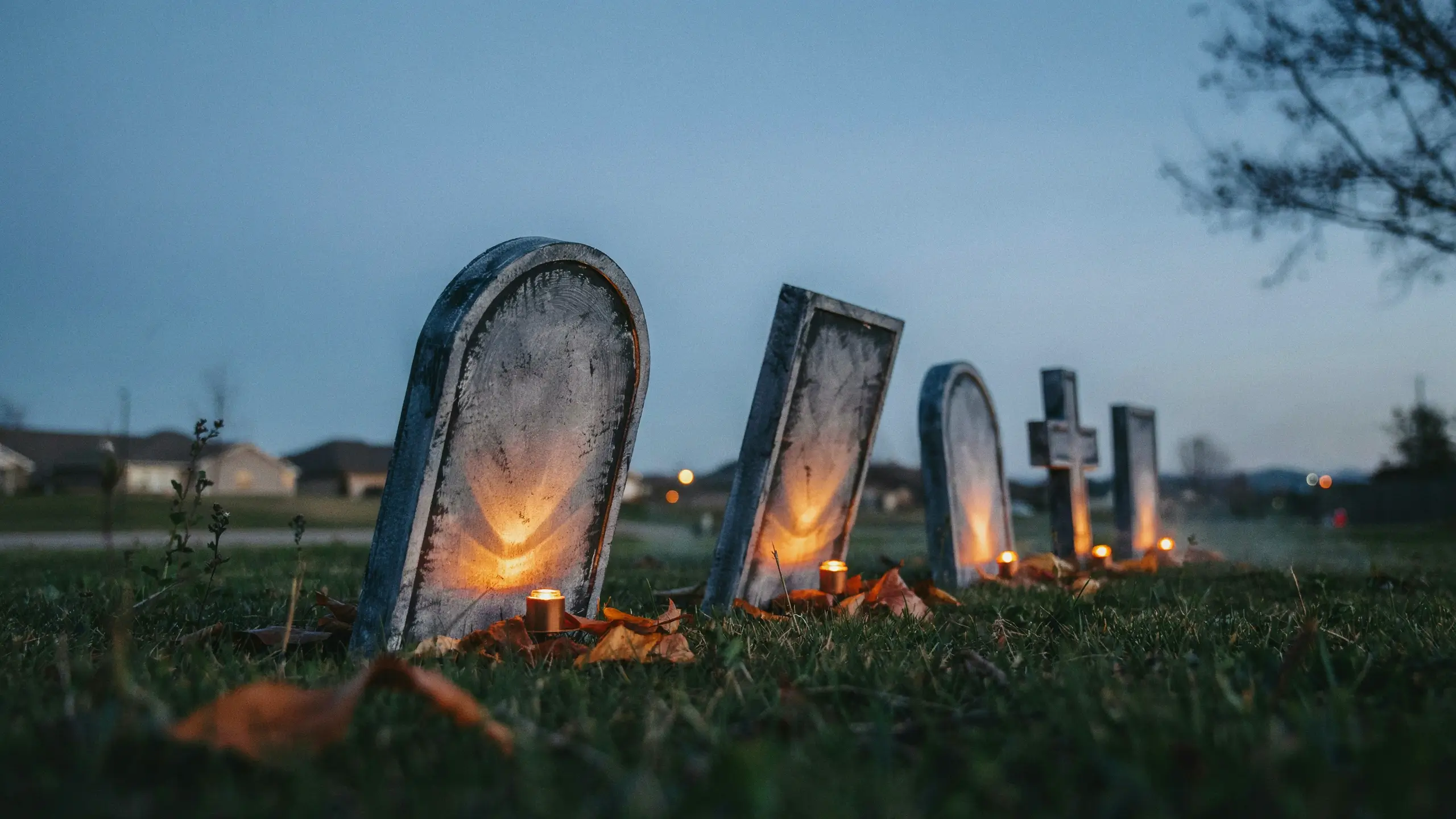 A row of DIY foam board tombstones and a cross acting as a Halloween graveyard decoration in a grassy suburban yard during twilight, illuminated by lit votive candles scattered with fallen autumn leaves. 