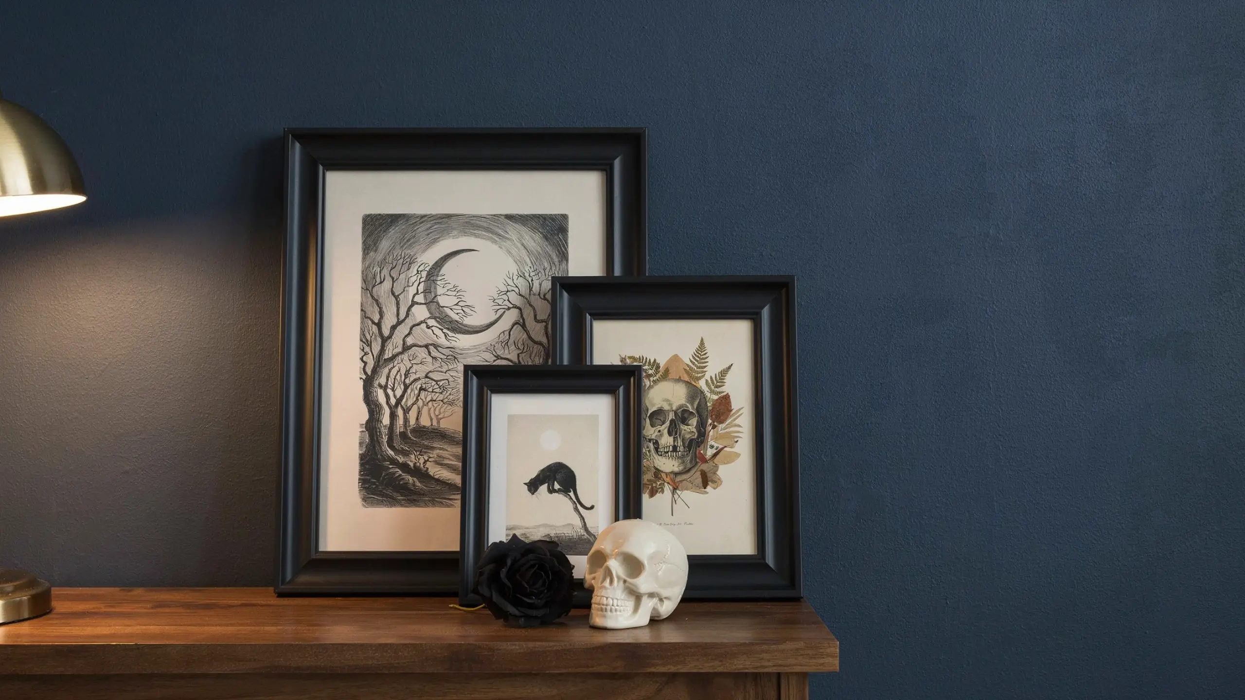 Three black framed DIY Halloween art prints featuring a crescent moon with bare winter trees, a black cat silhouette, and a vintage skull with autumn foliage, arranged in a cascading layout on a dark blue textured wall above a warm wooden shelf. A white decorative skull and a black artificial rose sit in the foreground under the glow of a brass reading lamp. 