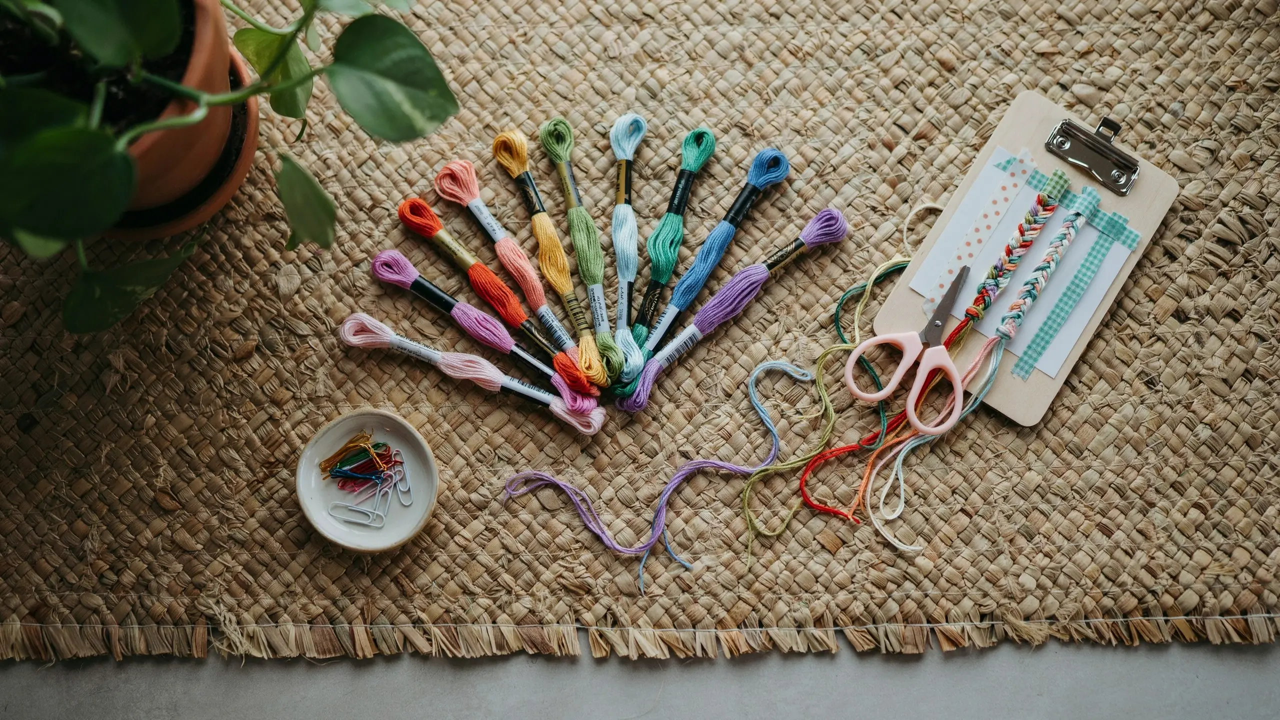 Overhead flat lay of vibrant embroidery floss threads, pink craft scissors, and a clipboard holding two partially braided friendship bracelets, arranged on a textured woven jute mat next to a potted plant.