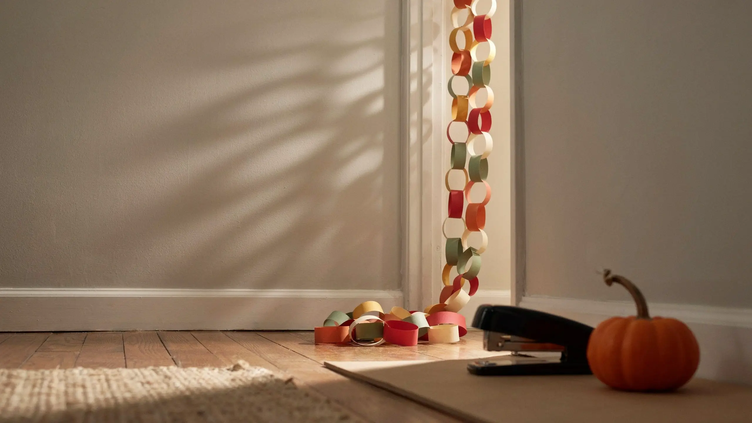 A colorful DIY gratitude paper chain countdown made of autumn-colored paper loops hanging from a door frame and pooling on a hardwood floor, placed next to a black stapler and a small decorative pumpkin in warm sunlight