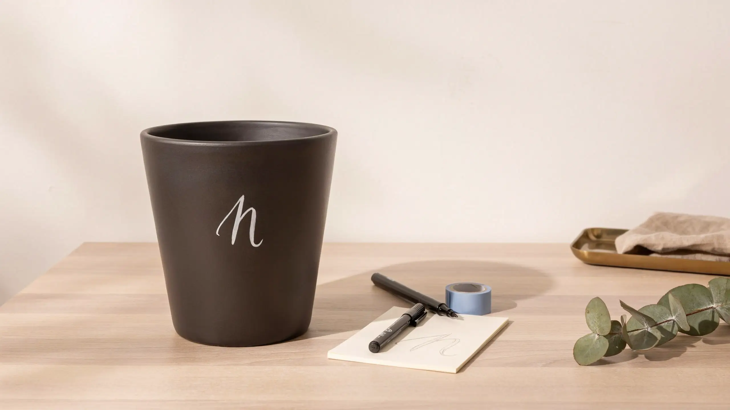 Matte black ceramic pot with white handwritten cursive "n" monogram on wooden table, surrounded by craft supplies and notepad for a DIY painted home decor project.