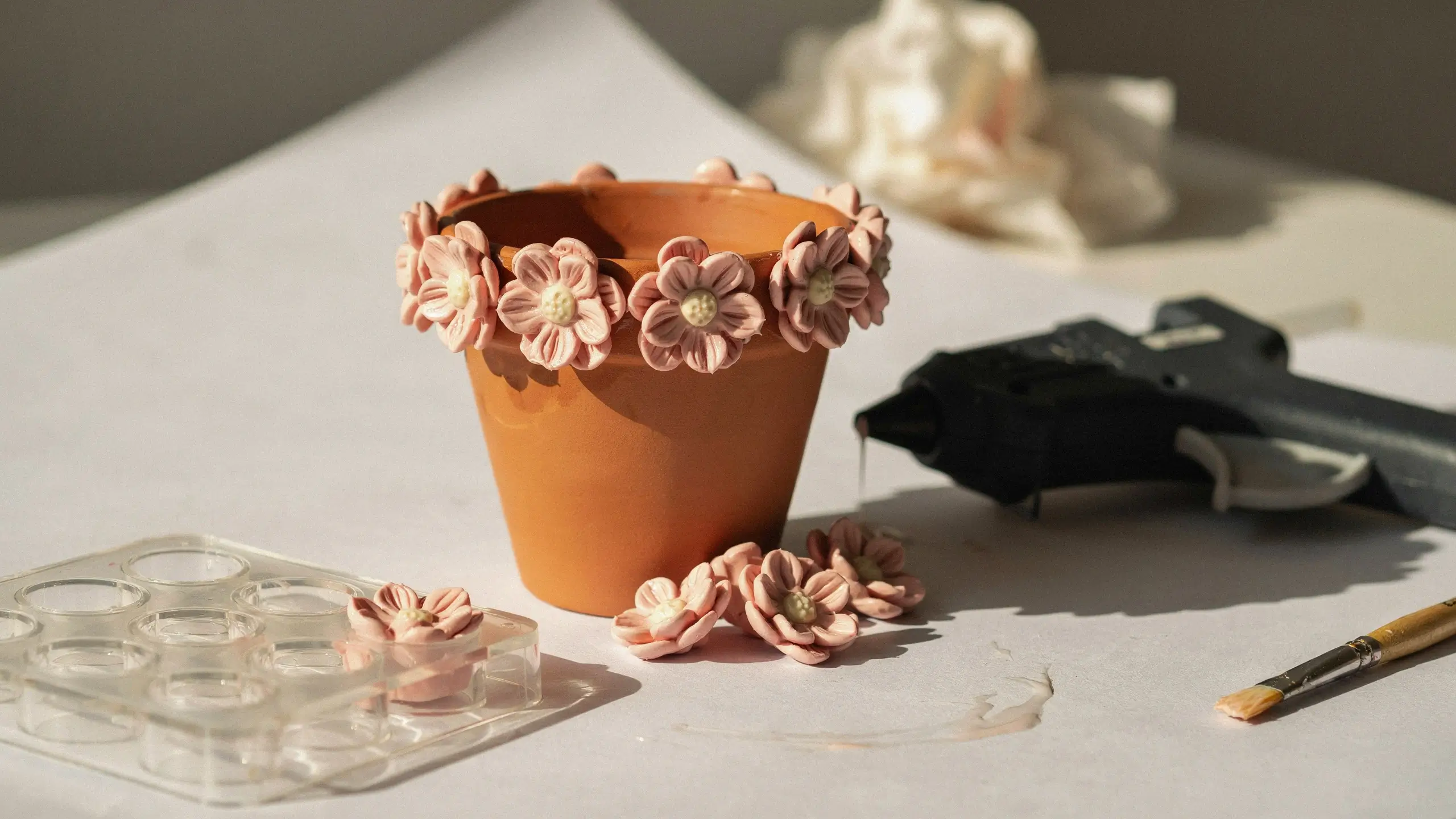 A terracotta pot decorated with a rim of pale pink 3D flowers, surrounded by crafting supplies including a black hot glue gun, a clear plastic molding tray, and a paintbrush on a white table.