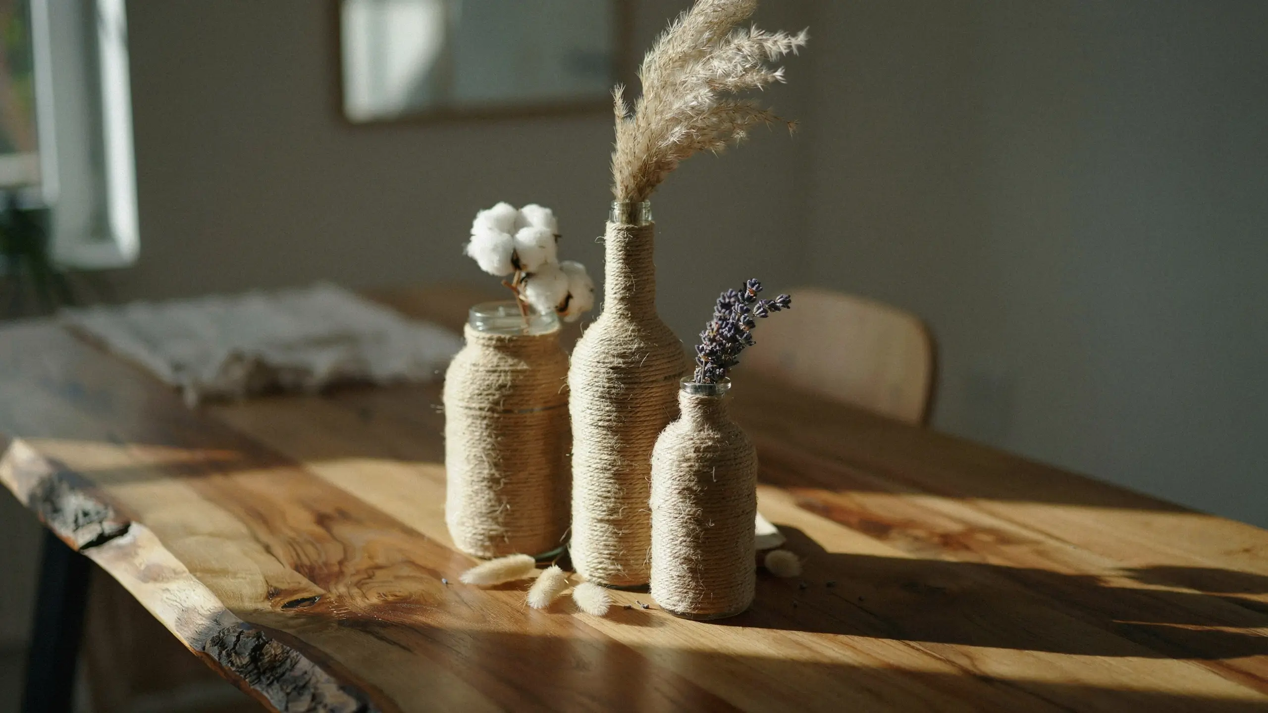 Three rustic glass bottles tightly wrapped in natural jute twine, repurposed as handmade vases on a sunlit wooden table. The arrangement features dried cotton bolls, tall pampas grass, and lavender sprigs, creating a boho-farmhouse aesthetic.