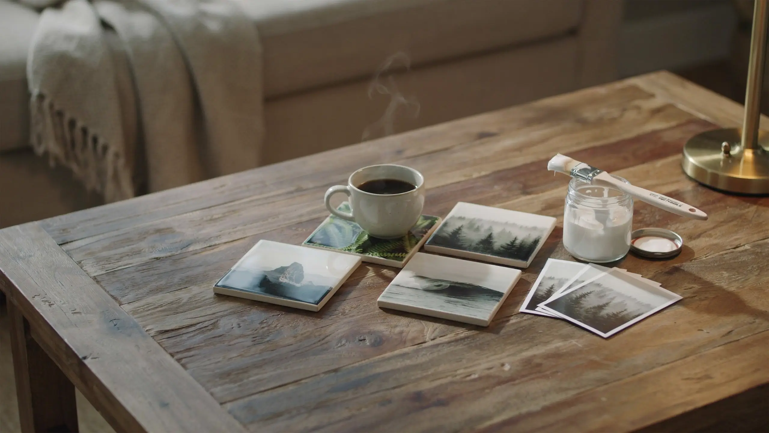  A rustic wooden table displaying four finished DIY Mod Podge photo coasters featuring landscape and botanical prints, accompanied by a steaming coffee mug, a white glue jar, and a paintbrush.