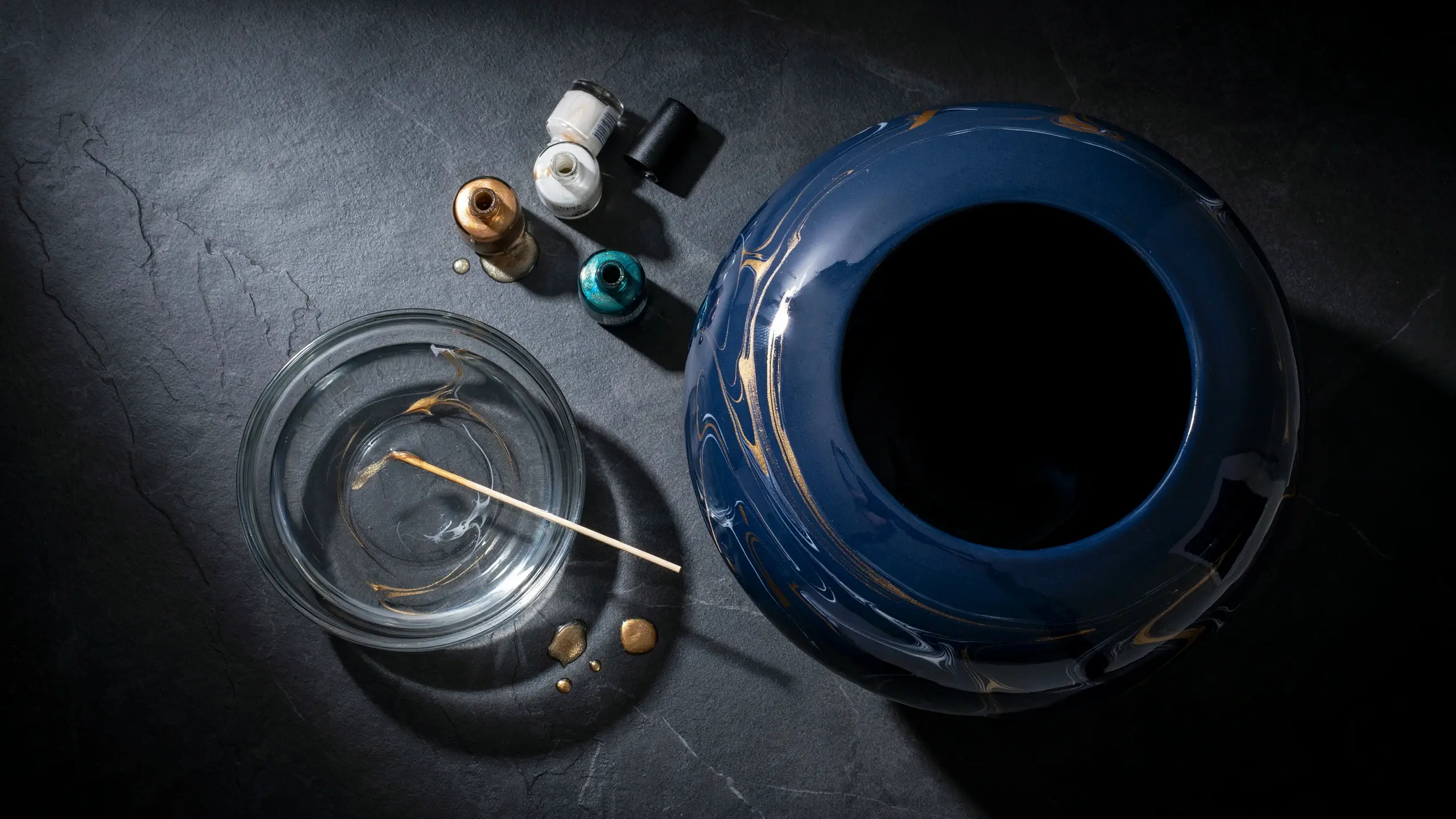 Overhead flat-lay of a DIY nail polish water marbling project for a dark blue ceramic pot, featuring a glass bowl with swirling paint and a wooden stirrer alongside bottles of gold, white, and teal polish