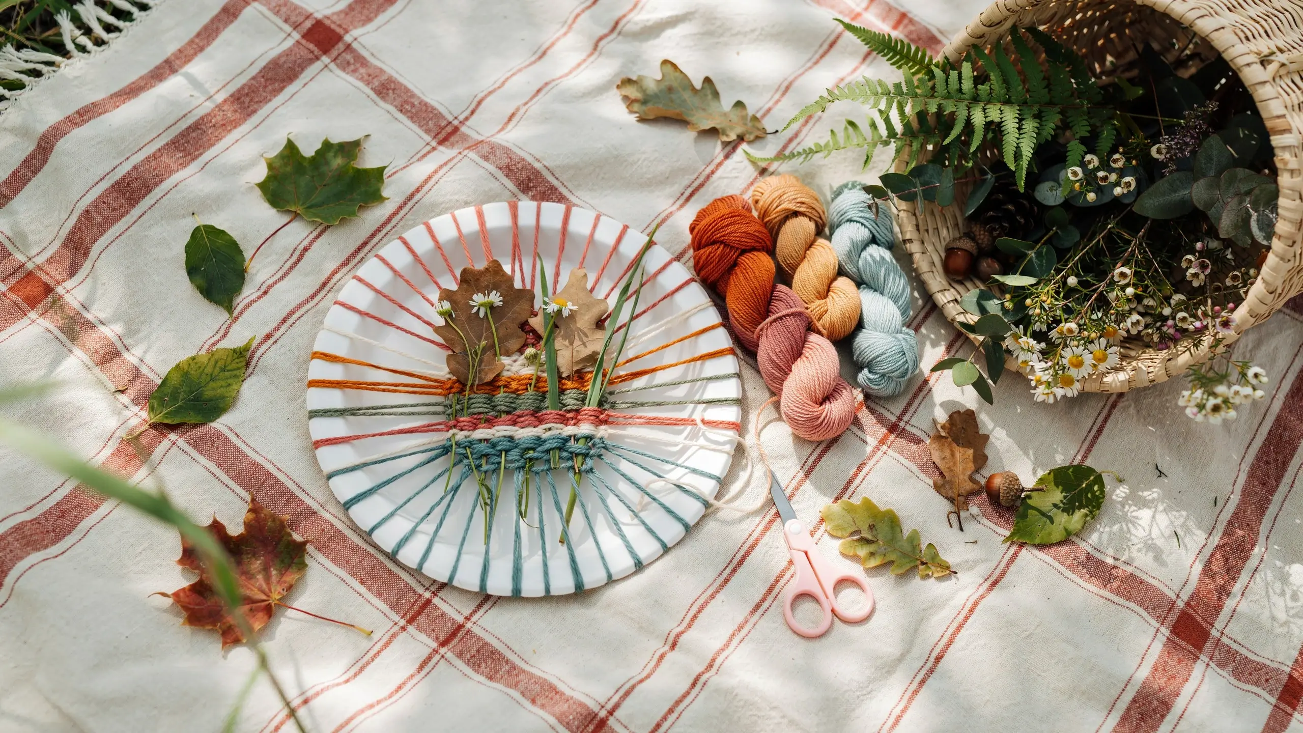An overhead close-up of a DIY nature weaving craft on a red and white plaid picnic blanket. A paper plate acts as a loom, woven with teal, orange, and pink yarn, holding fresh white daisies, green grass, and dried oak leaves. Beside it sit skeins of yarn, pink-handled scissors, and a wicker basket brimming with fern fronds and botanical floral elements.