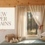 How We Finally Fixed Our Camper’s Ugly Curtains (No Sewing Machine Required) Beige no-sew camper curtains tied back with a red ribbon on an RV window, set within a cozy interior featuring an open book, a coffee mug, and a greening potted plant on a wooden shelf.