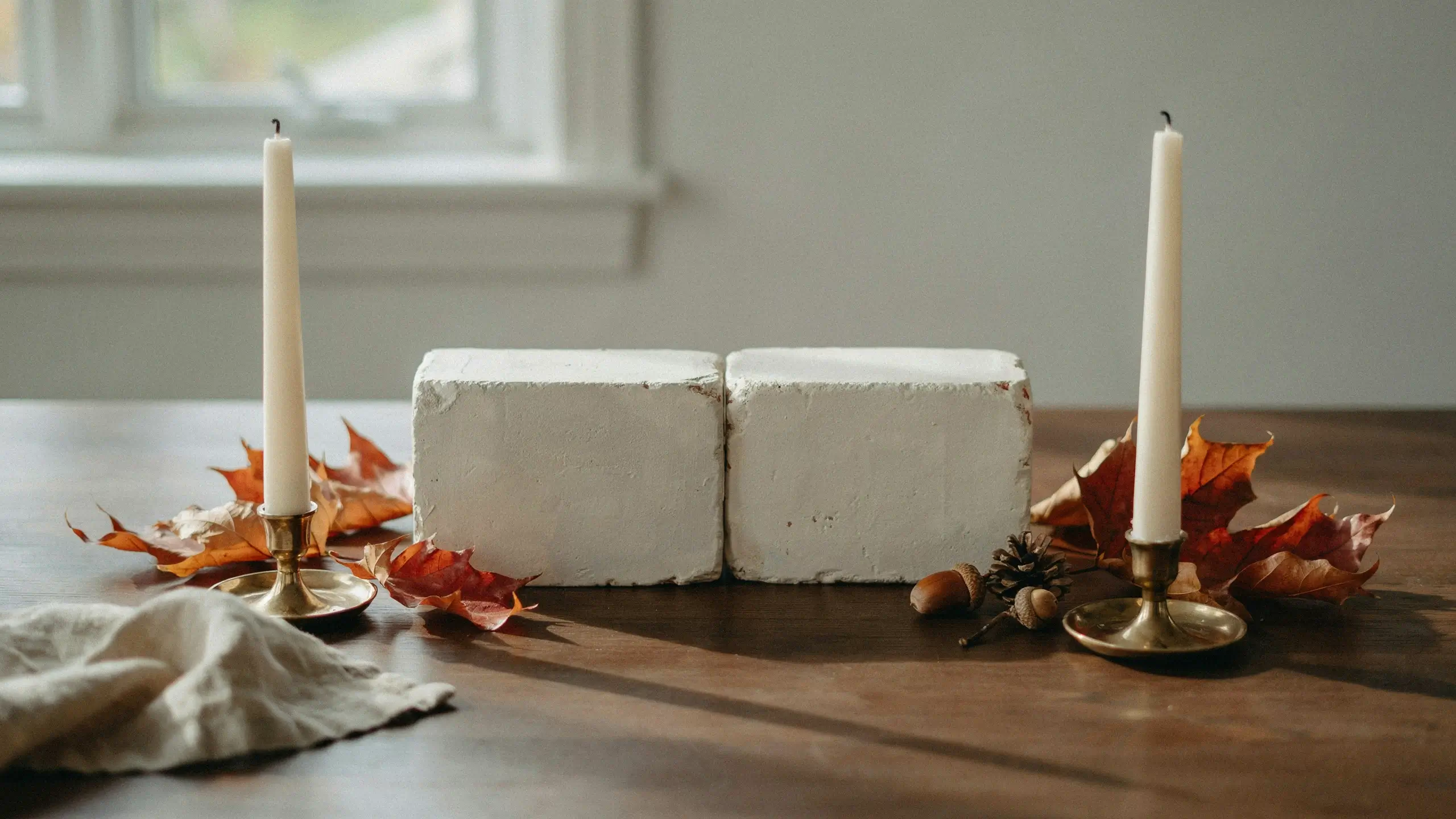 Two white painted brick "Give Thanks" blocks styled with white taper candles, autumn maple leaves, acorns, and a pinecone on a rustic wooden table.