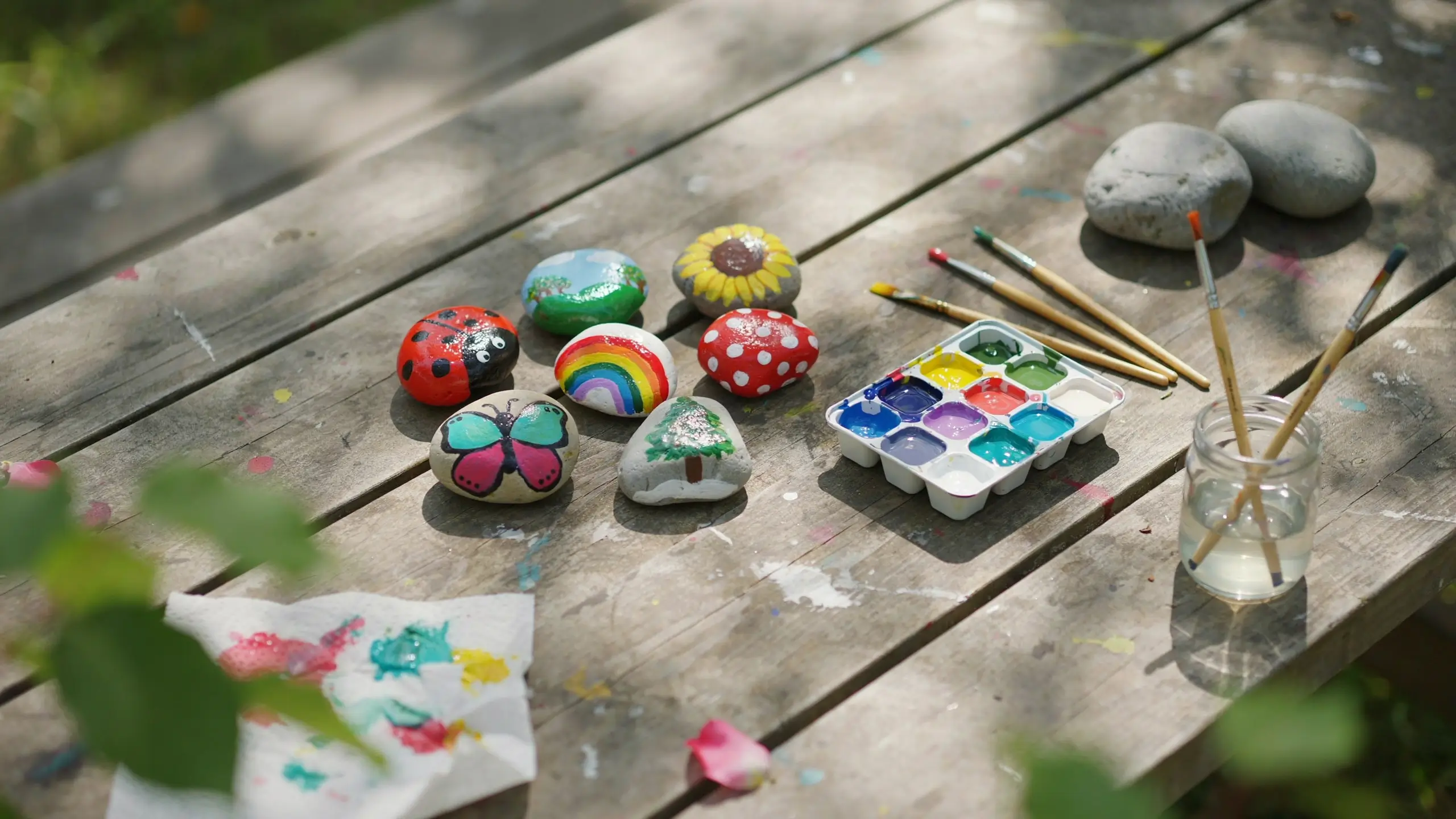 A rustic wooden table showcasing a collection of colorful, hand-painted rocks featuring a ladybug, butterfly, rainbow, sunflower, and a tree, surrounded by an open watercolor palette, paintbrushes, a glass jar with water, and a paper towel with paint splatters during an outdoor summer craft session.