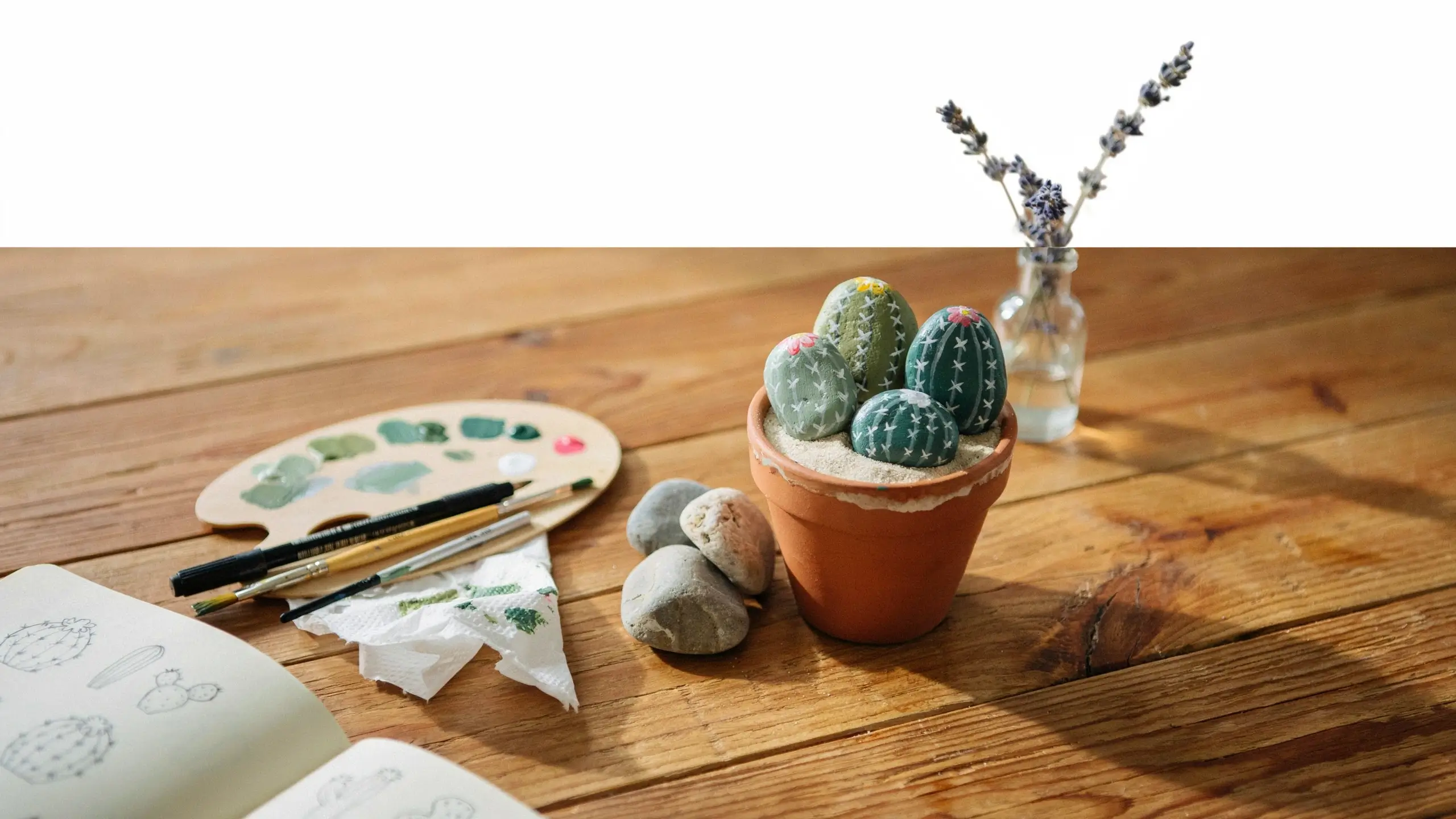 Hand-painted stone cacti in a terracotta pot with white sand, surrounded by a green paint palette, brushes, and a sketchbook on a rustic wooden table.