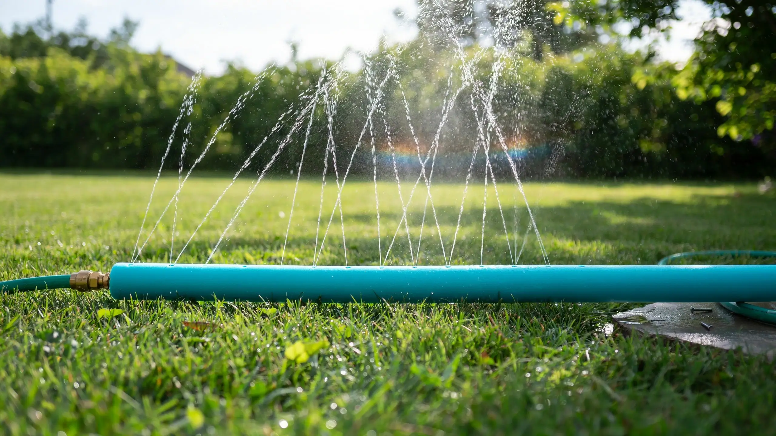 Teal DIY pool noodle sprinkler connected to a garden hose spraying water arcs onto a green lawn.
