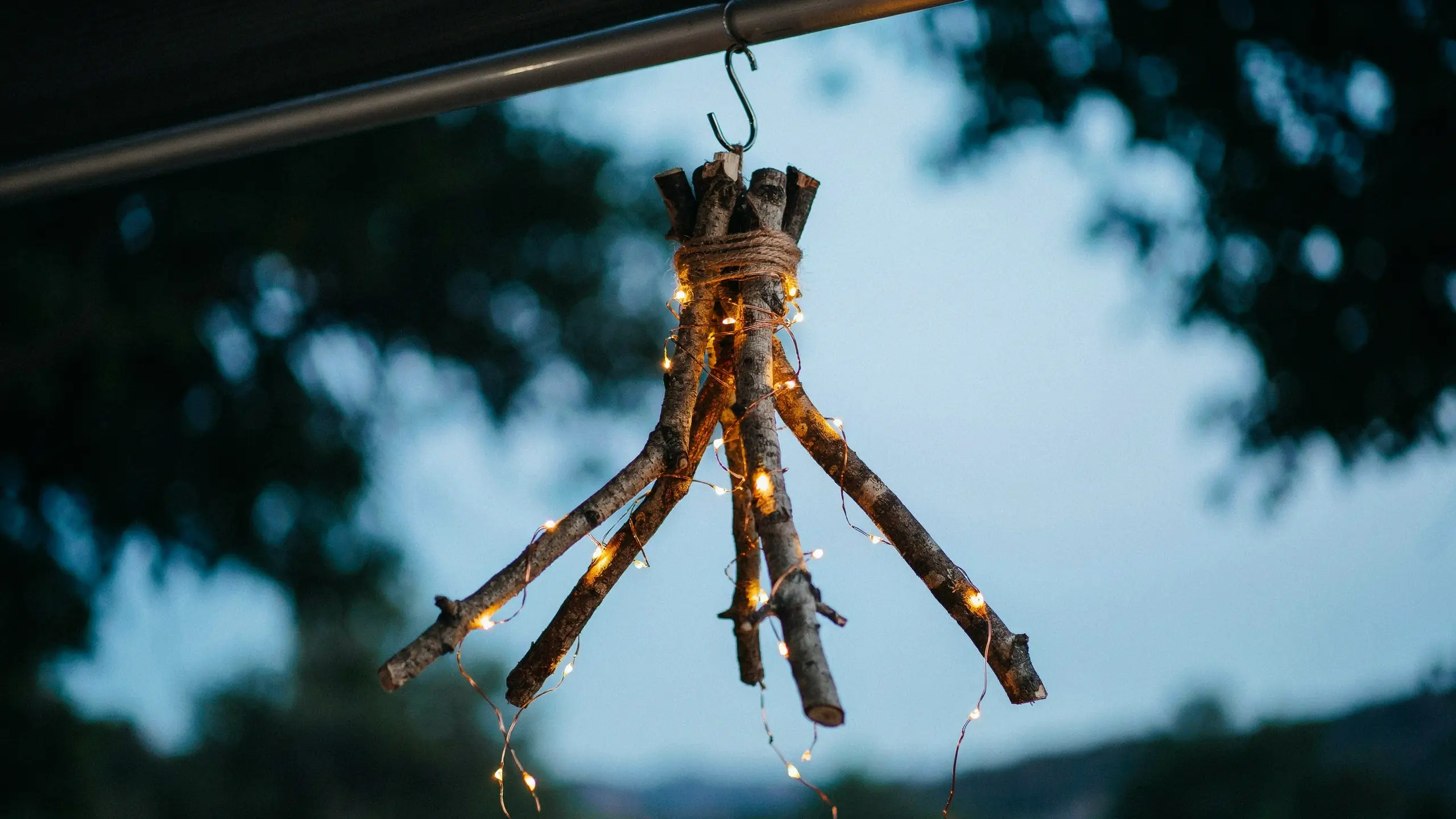 Rustic DIY branch chandelier wrapped in jute twine and illuminated by warm fairy lights hanging outdoors at dusk. 