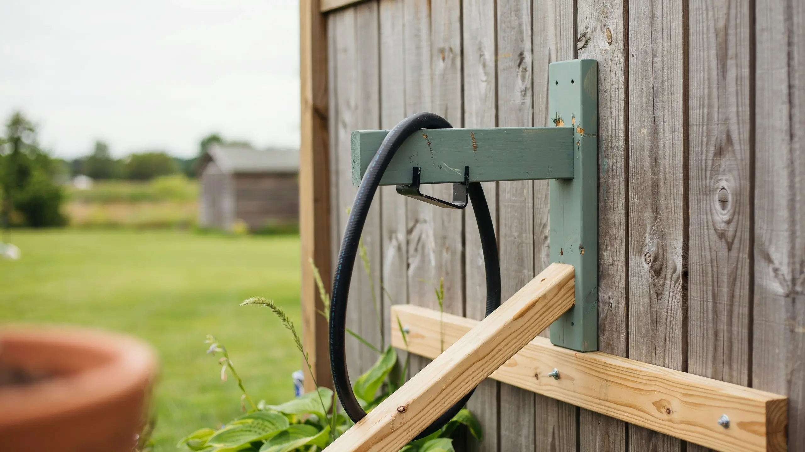 Sage green painted wooden wall-mounted garden hose holder with black metal hook installed on a weathered vertical wood fence.