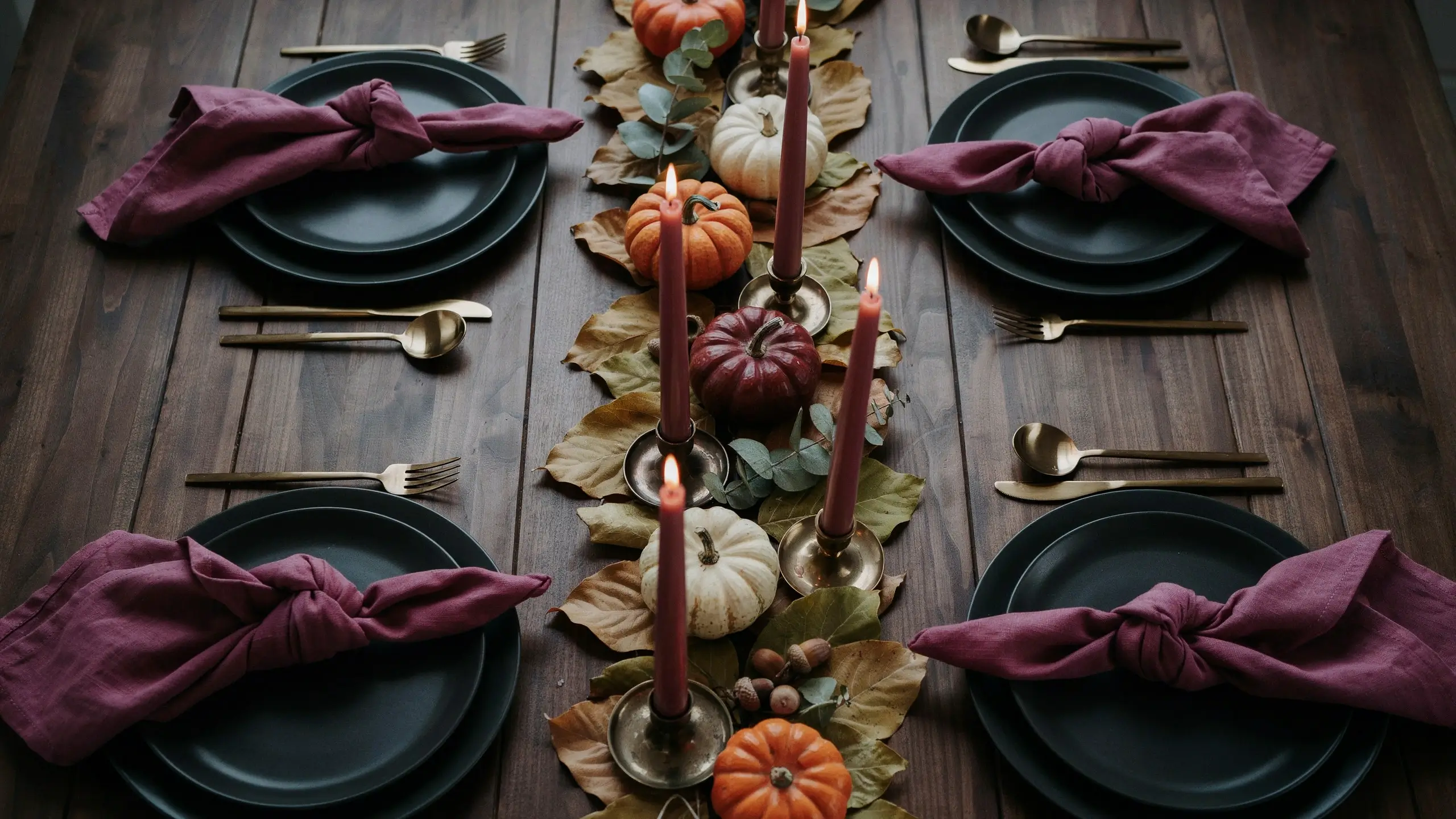 Moody autumn tablescape featuring dark plates, burgundy knotted napkins, and brass flatware surrounding a rustic centerpiece of pumpkins and dried leaves, designed as the perfect staging area for displaying handmade DIY skull candlesticks. 